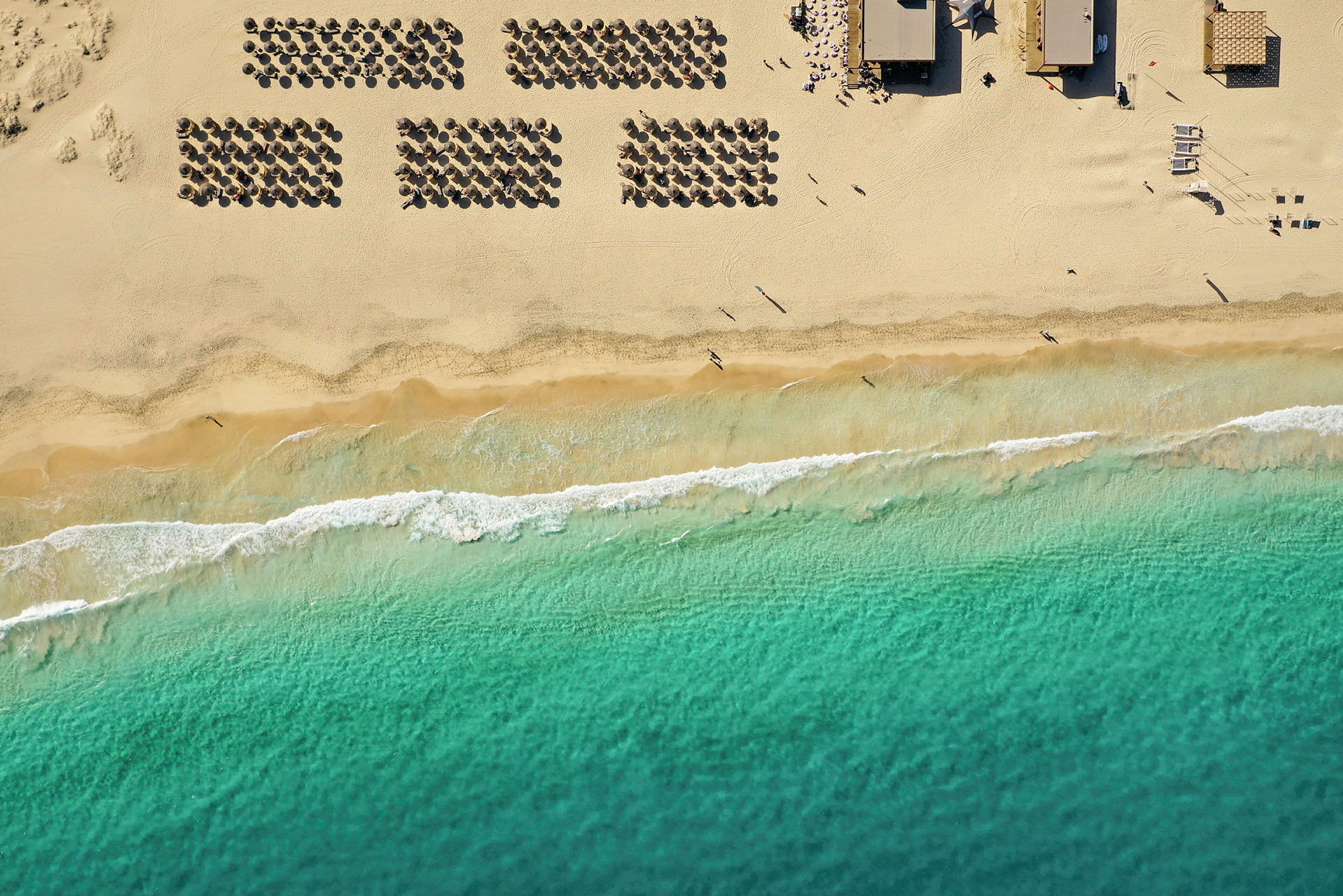 Flyfoto av en strand med parasoller, sand og havbølger. (AI-generert tekst)