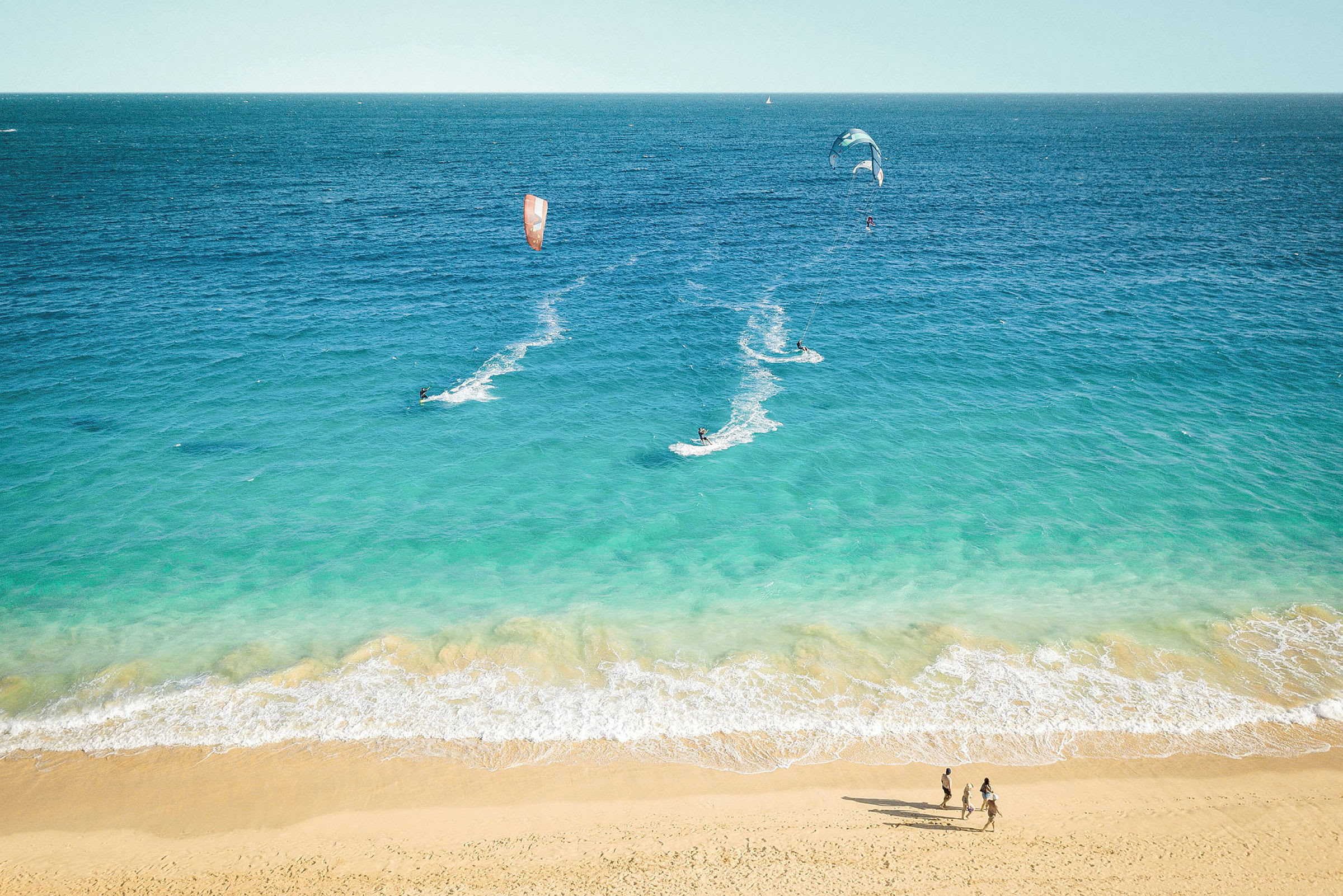 Strand med blått hav hvor personer driver med kitesurfing og går tur. (AI-generert tekst)