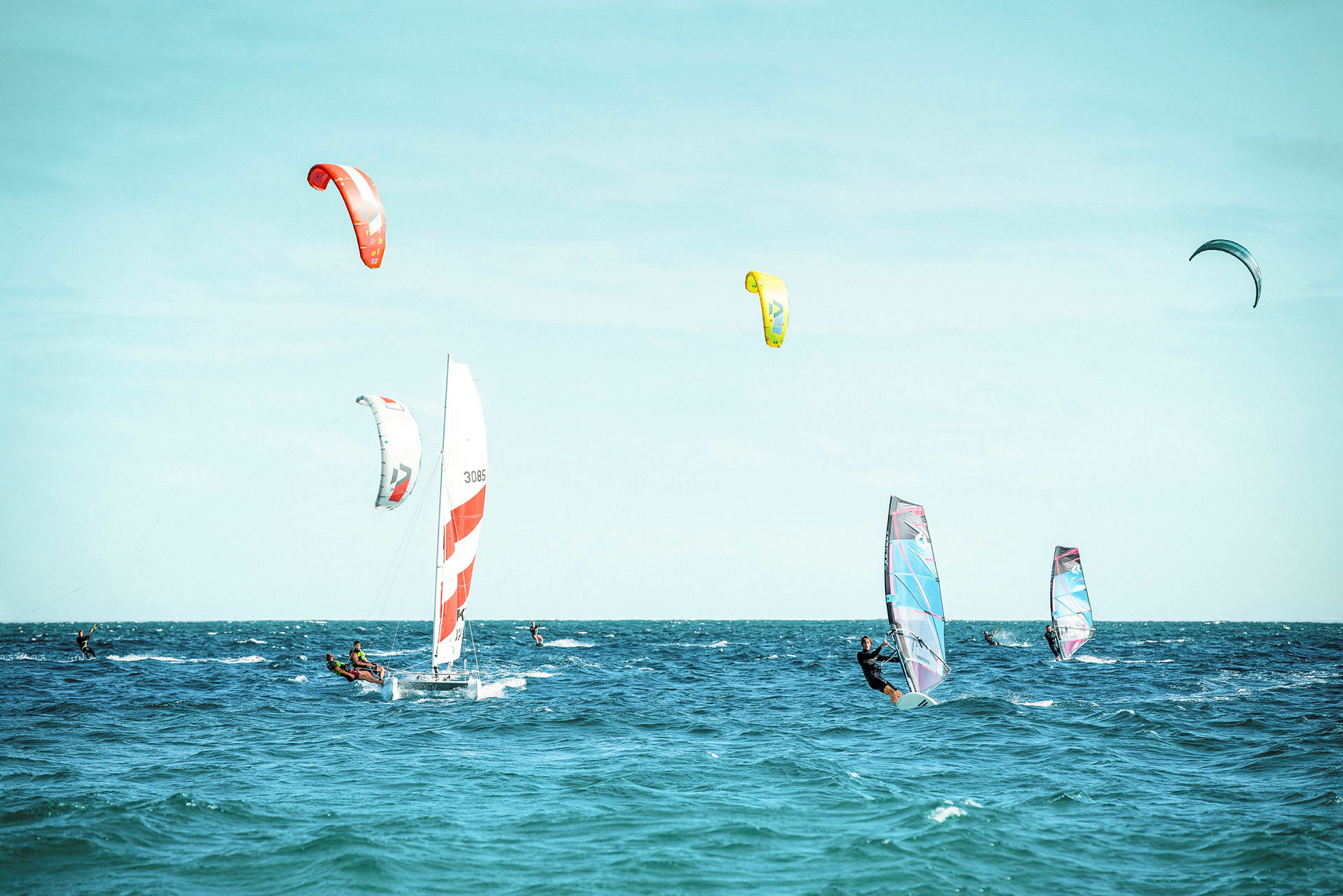Flere personer deltar i forskjellige vindsportaktiviteter på havet, med seilbåt, kitesurfing og seilbrett. (AI-generert tekst)