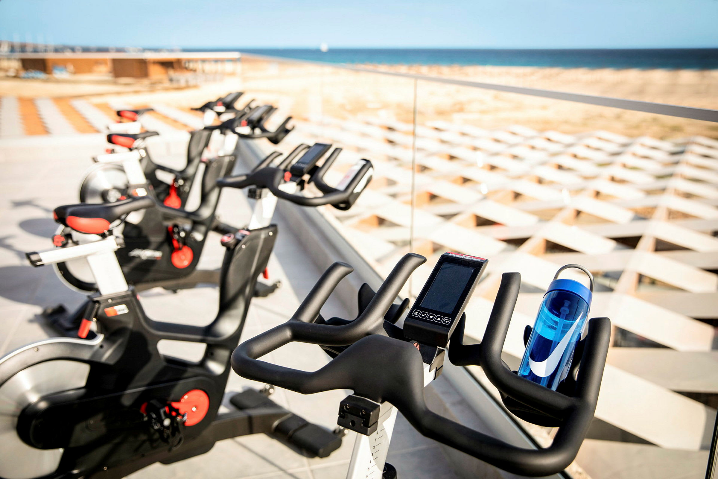 Bilde av flere treningssykler plassert ved et glassrekkverk med utsikt over stranden og havet. (AI-generert tekst)