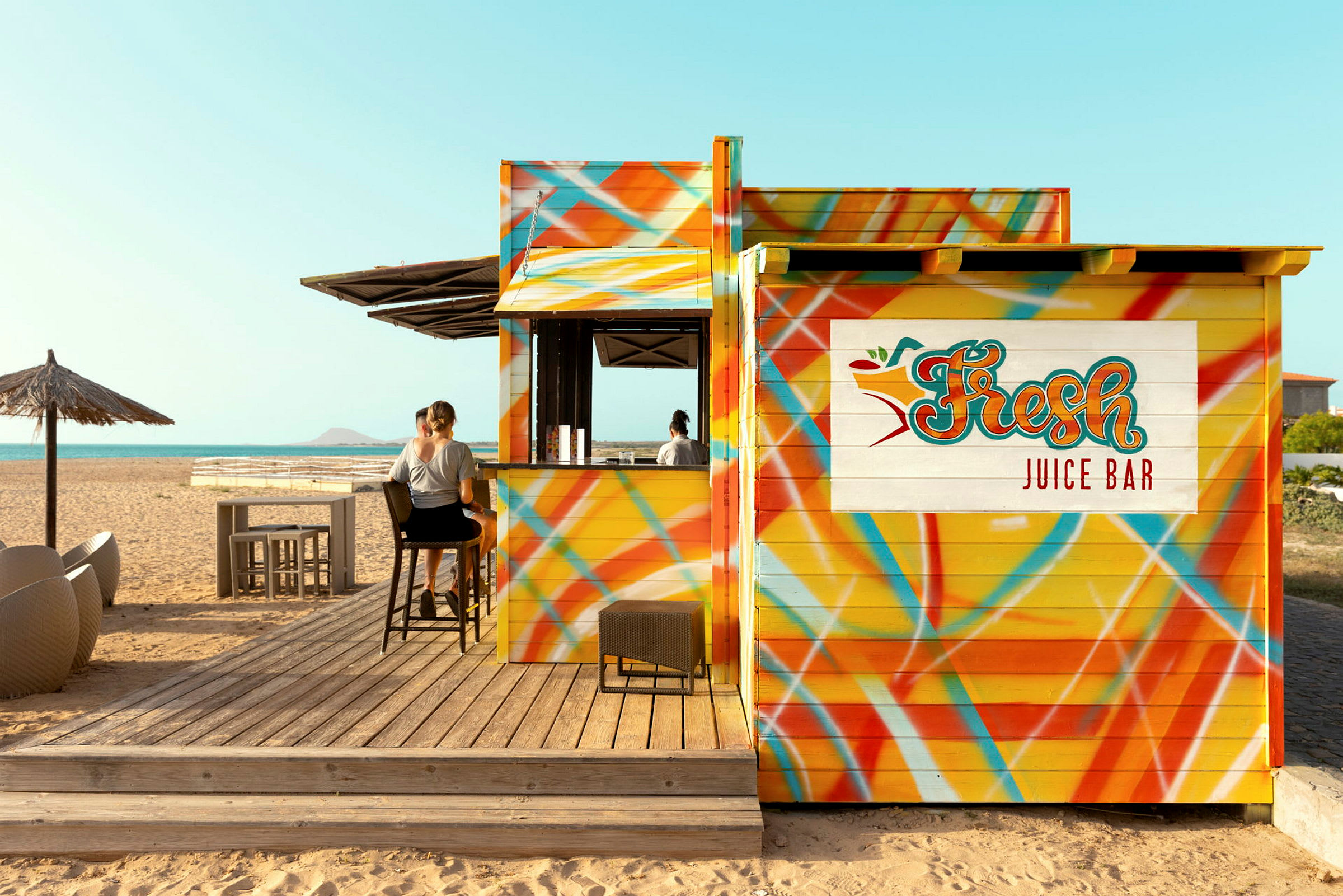 En fargerik juicebar på stranden med personer som sitter på barkrakker. (AI-generert tekst)