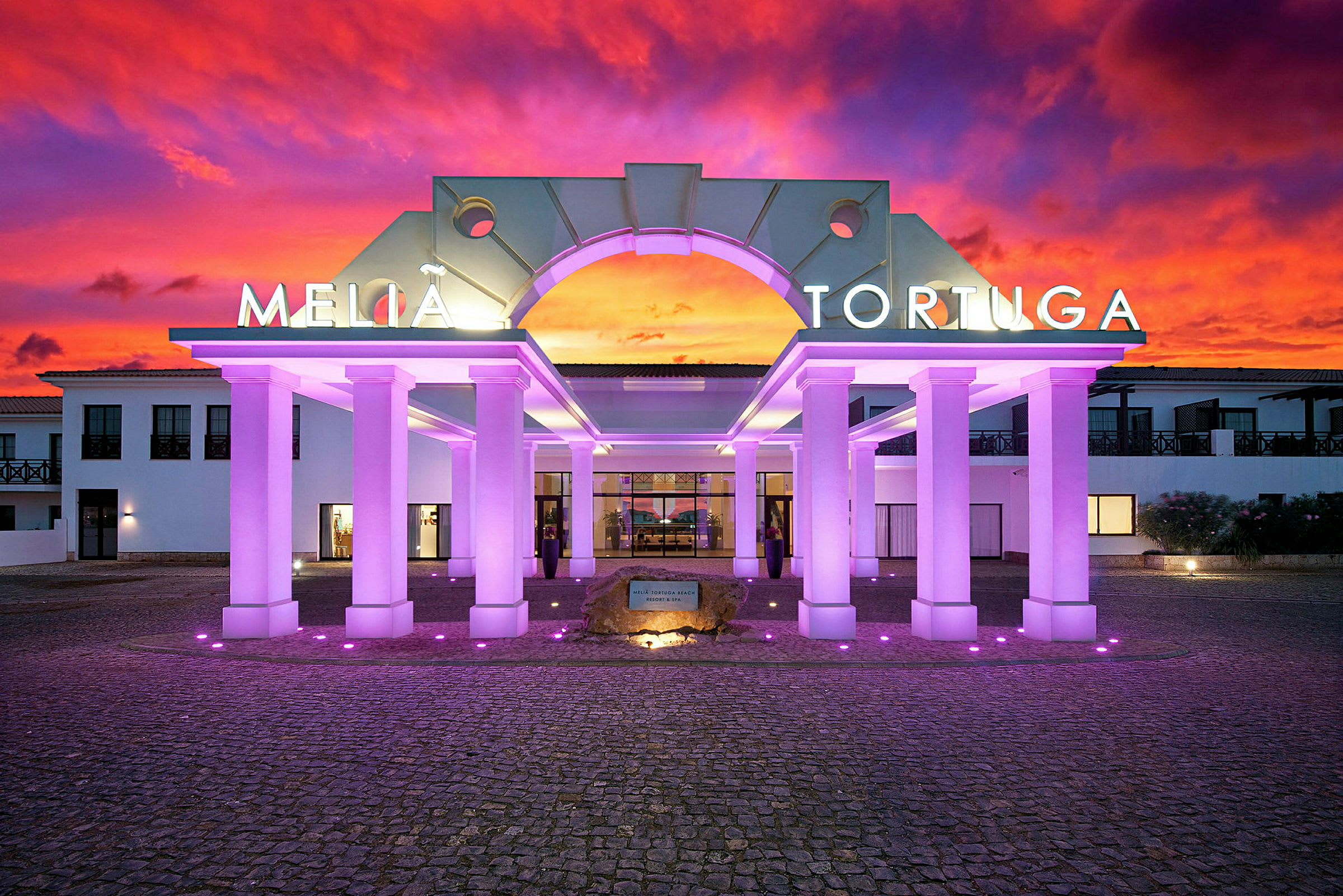Inngangen til hotellet med opplyste søyler og en fargerik solnedgang i bakgrunnen. (AI-generert tekst)