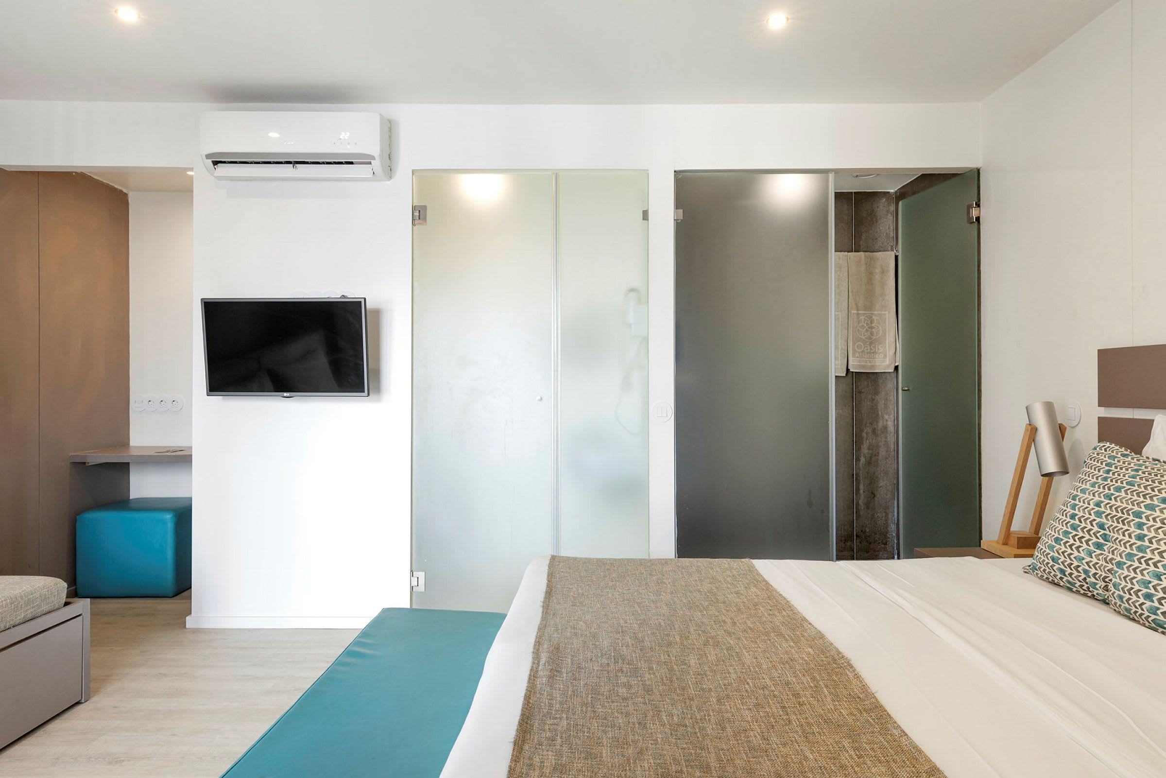 Et moderne hotellrom med en seng, TV og en arbeidsplass. Glassdører fører til badet. (AI-generert tekst)