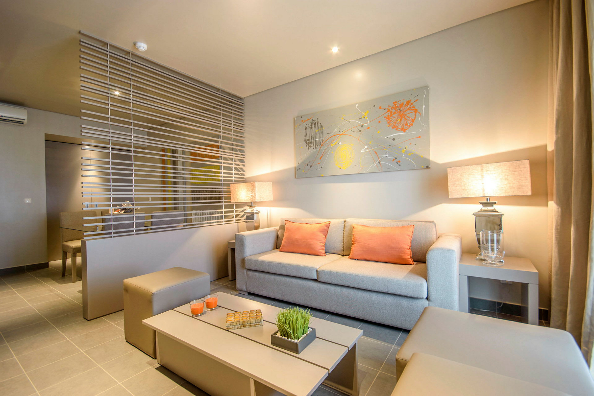 Stue med moderne innredning, beige sofa og oransje puter, adskilt med dekorativt horisontalt nett. (AI-generert tekst)