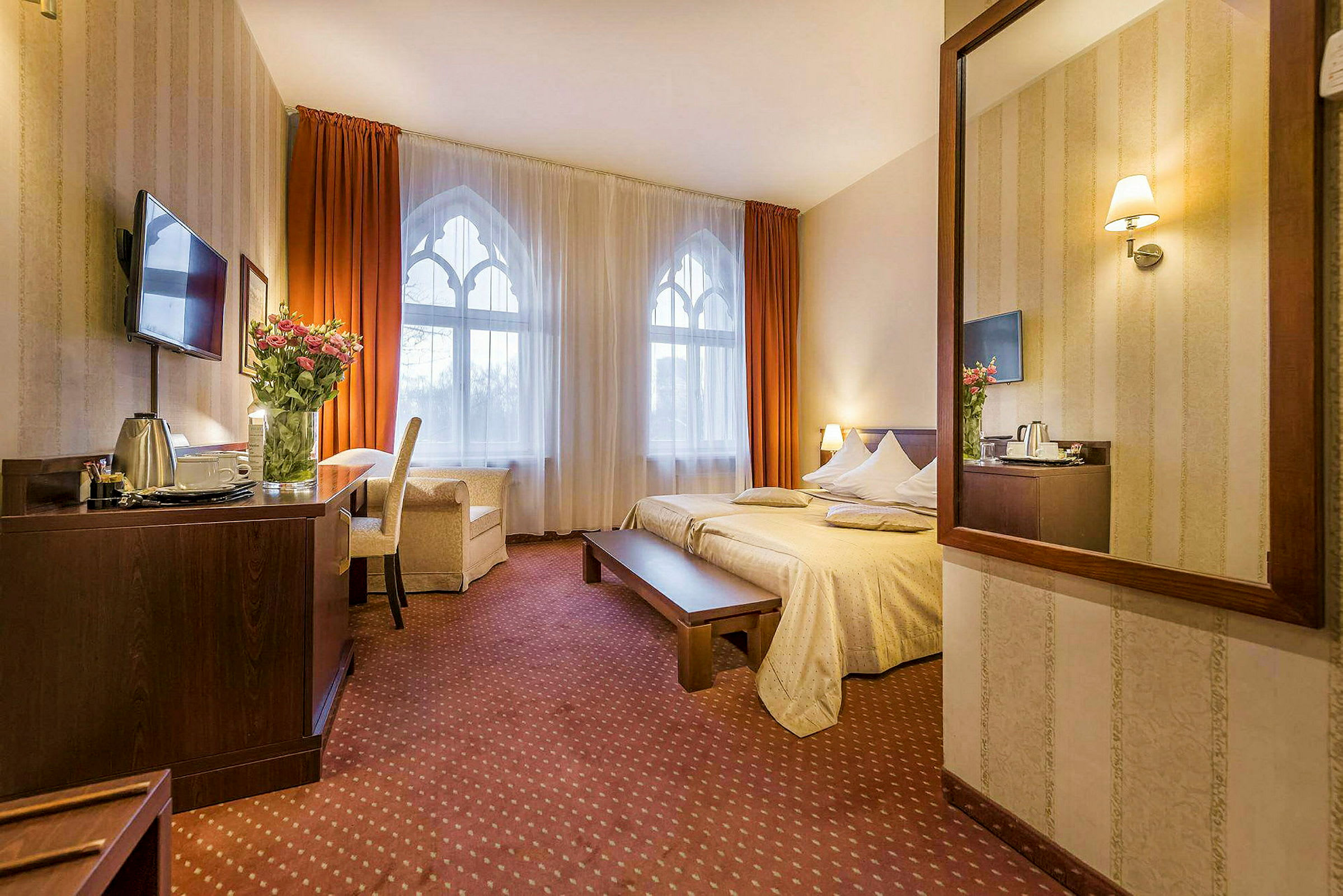 Et hotellrom med en stor seng, et bord og en stol. Vinduet har hvite gardiner med oransje draperier. (AI-generert tekst)