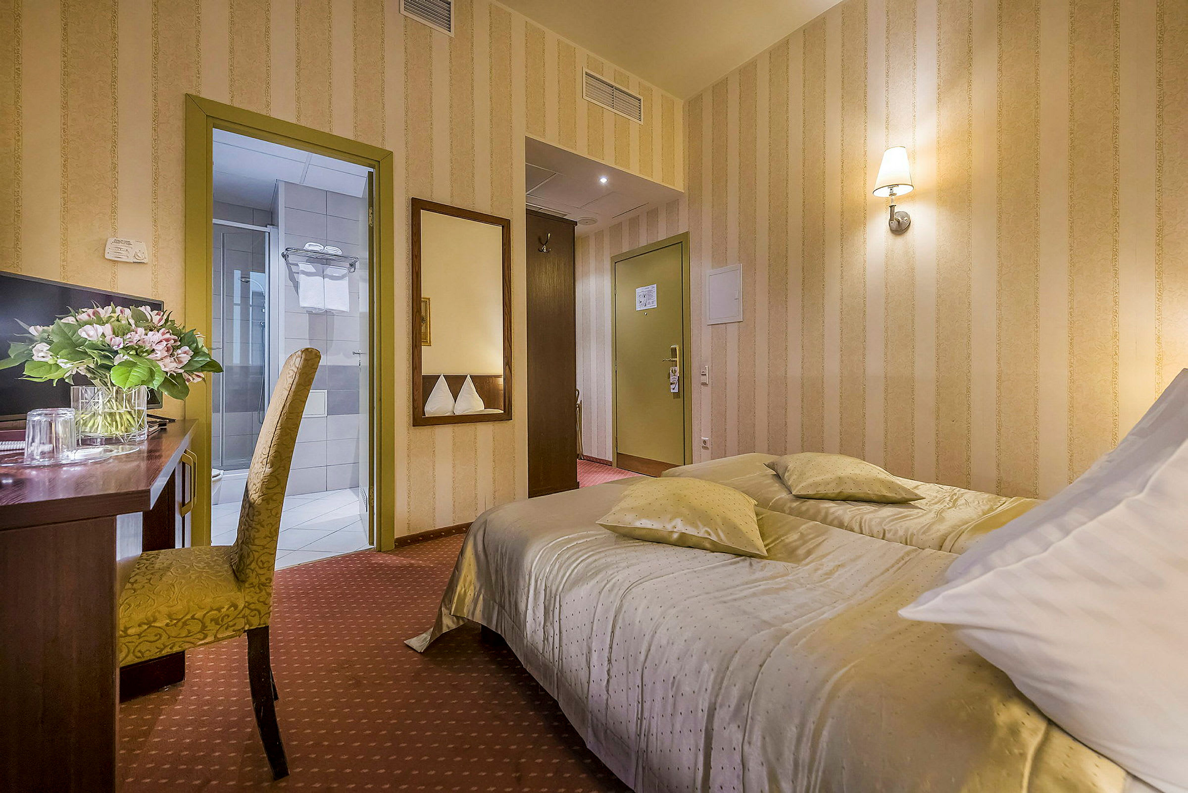 Et hotellrom med en oppredd seng, et skrivebord med en blomsterkrans og en stol samt dører til bad og gang. (AI-generert tekst)