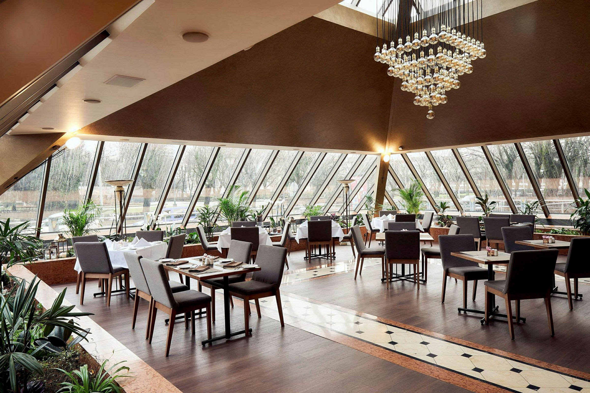 En elegant restaurant med store vinduer, mange bord og stoler, samt lysekrone i taket. (AI-generert tekst)