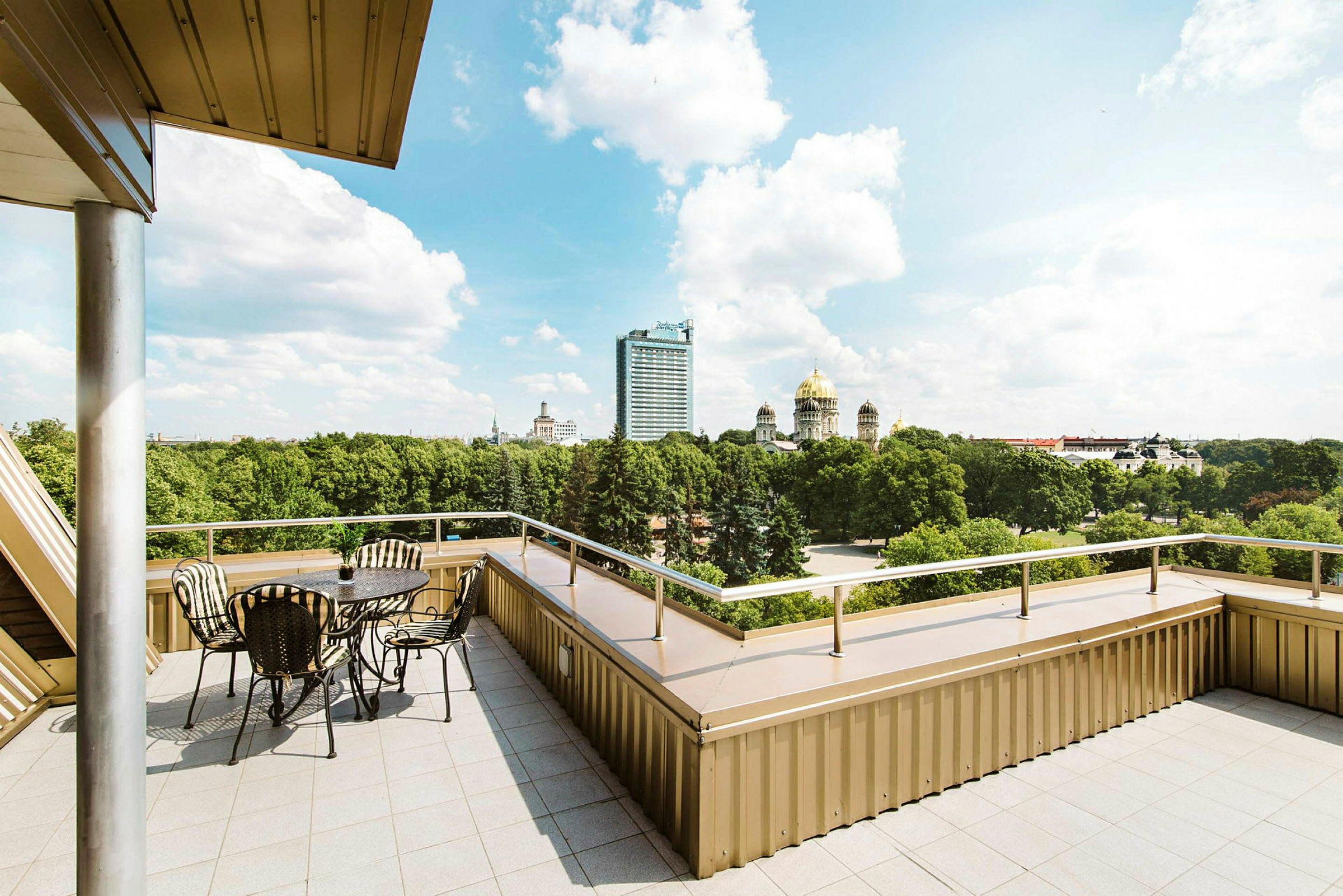 En terrasse med bord og stoler samt utsikt over en park og flere bygninger, inkludert en med gullkuppel. (AI-generert tekst)