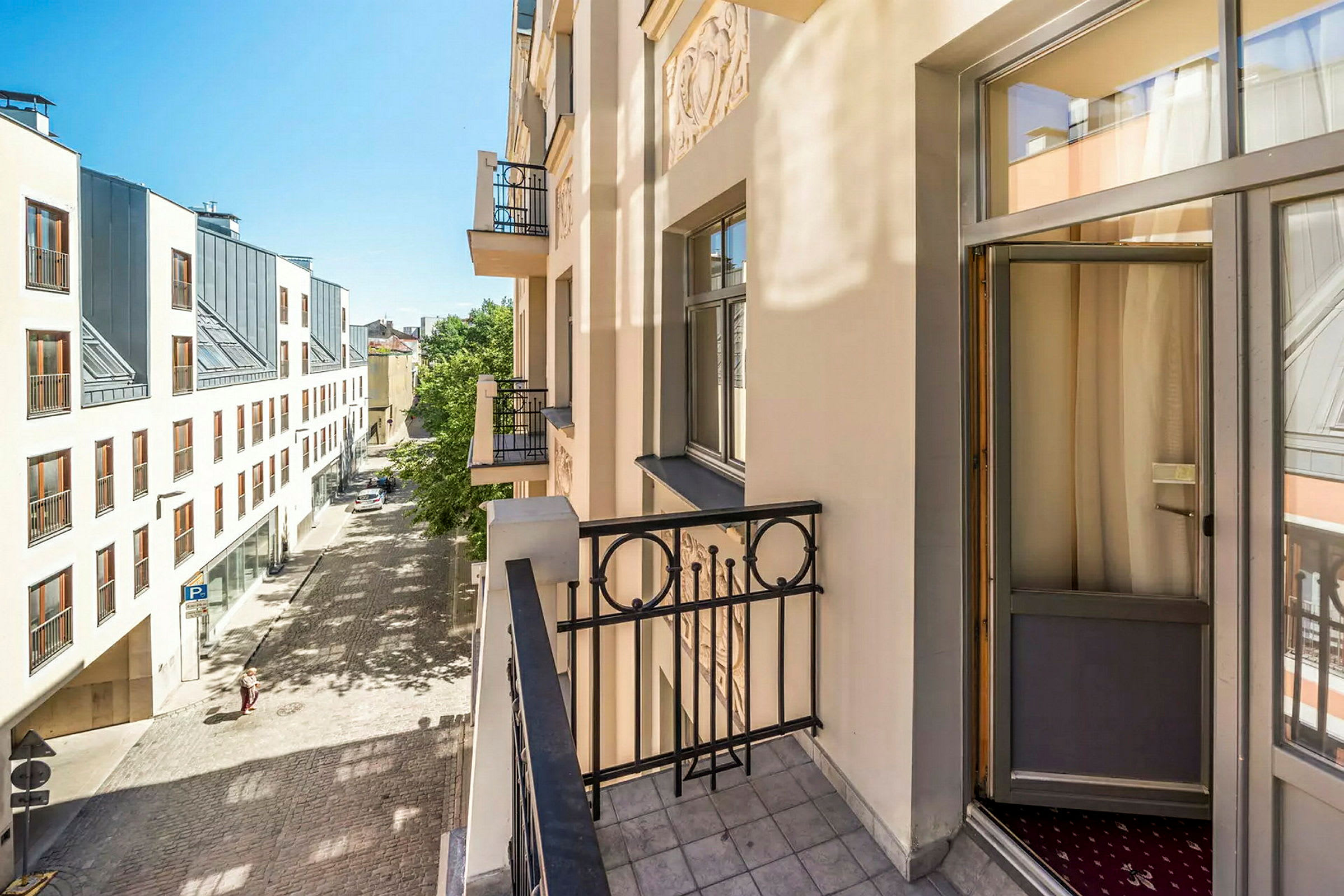 Bildet viser en balkong på en lys bygning med utsikt over en brosteinsgate og et moderne hus med store vinduer på den andre siden av gaten. (AI-generert tekst)