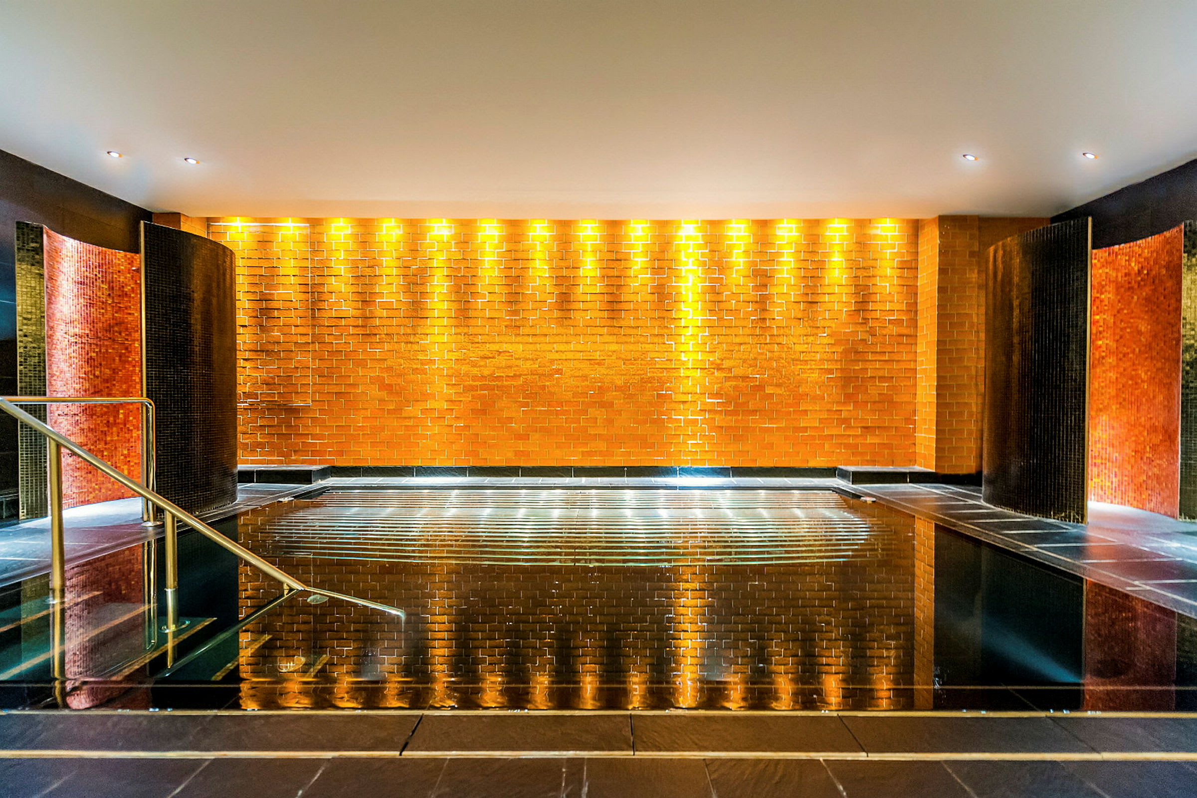 Et innendørs spa med et skinnende svart flislagt bassengområde, omgitt av en vegg med oransje fliser og belysning. (AI-generert tekst)