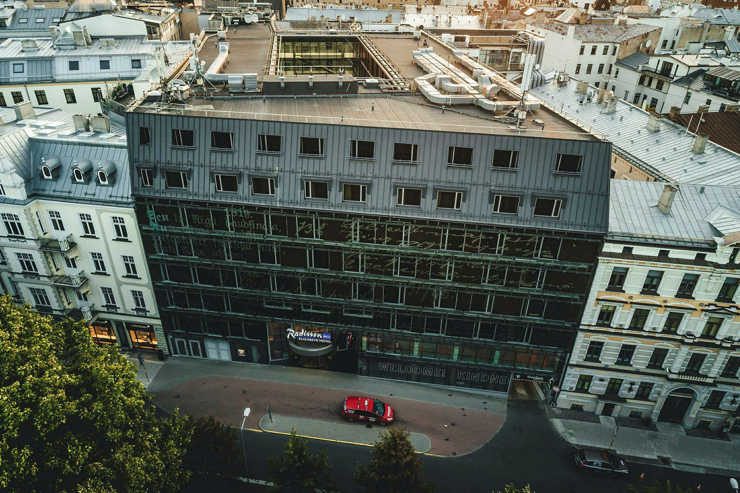 Luftfoto av en bygning med glassfasade og flere etasjer omgitt av andre bygninger. (AI-generert tekst)