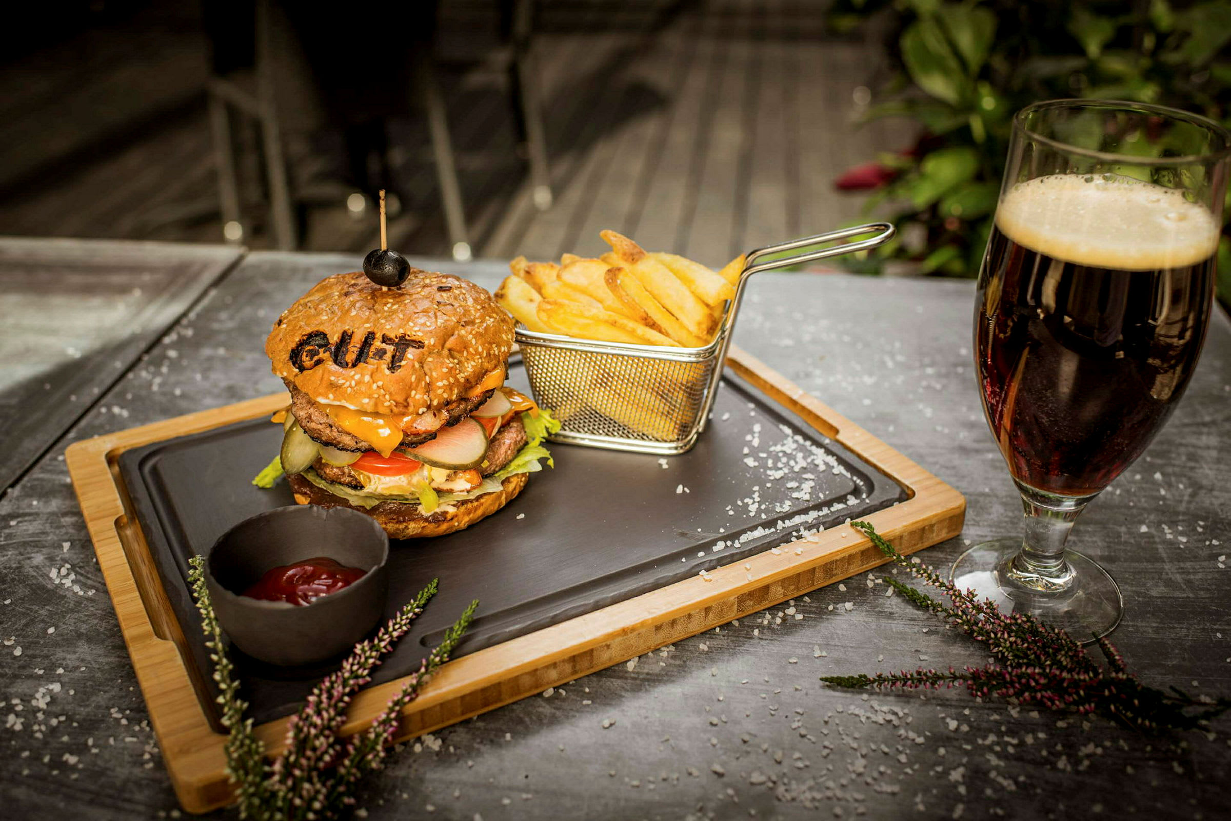 En tallerken med en hamburger og pommes frites samt en skål med ketchup, plassert på et bord ved siden av et glass mørk drikke. (AI-generert tekst)