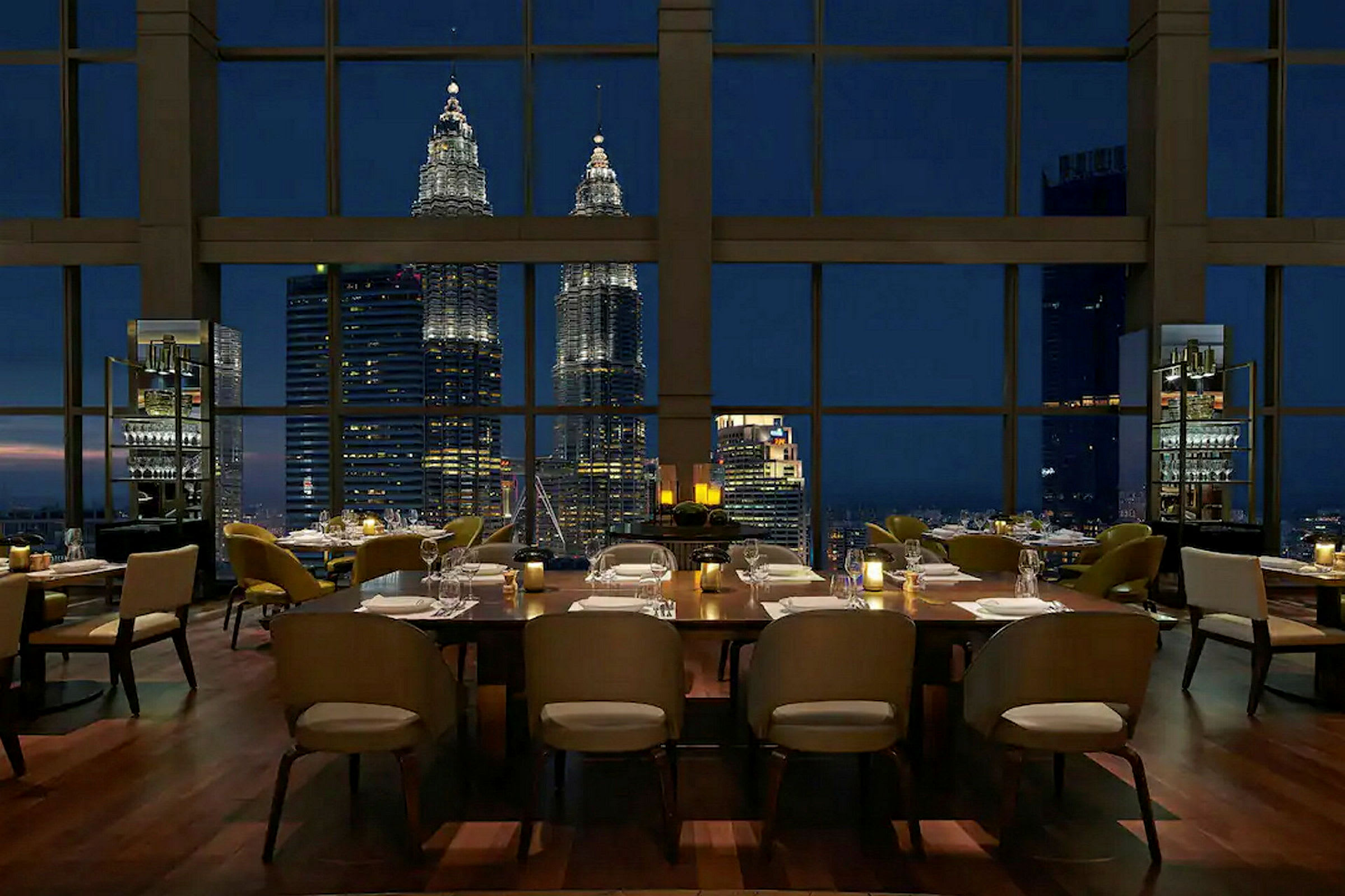 En restaurant med oppsatte bord som har utsikt mot Petronas Twin Towers gjennom store vinduer om kvelden. (AI-generert tekst)