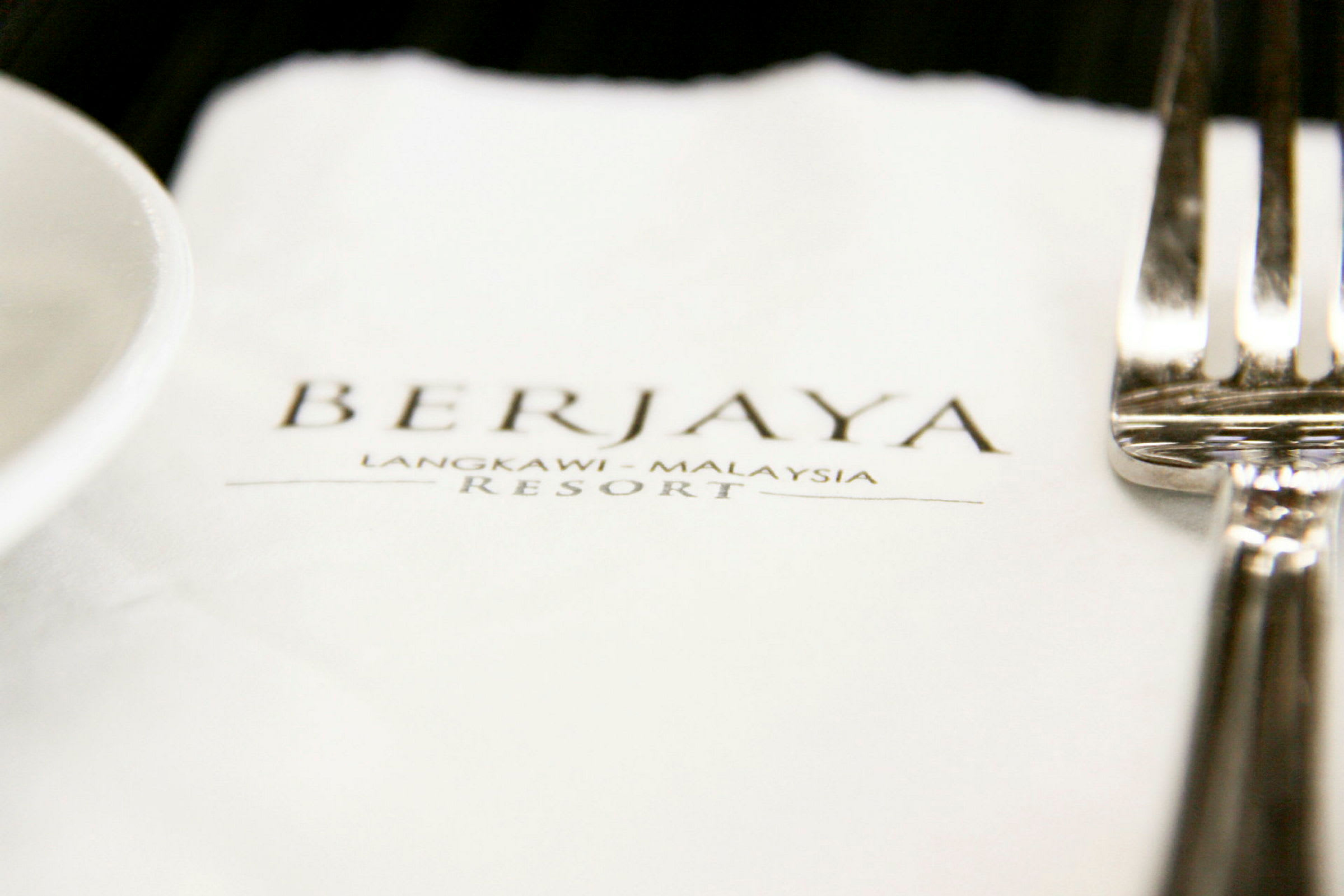 Et nærbilde av en serviett med teksten "BERJAYA Langkawi - Malaysia Resort" samt en gaffel og en del av en tallerken. (AI-generert tekst)