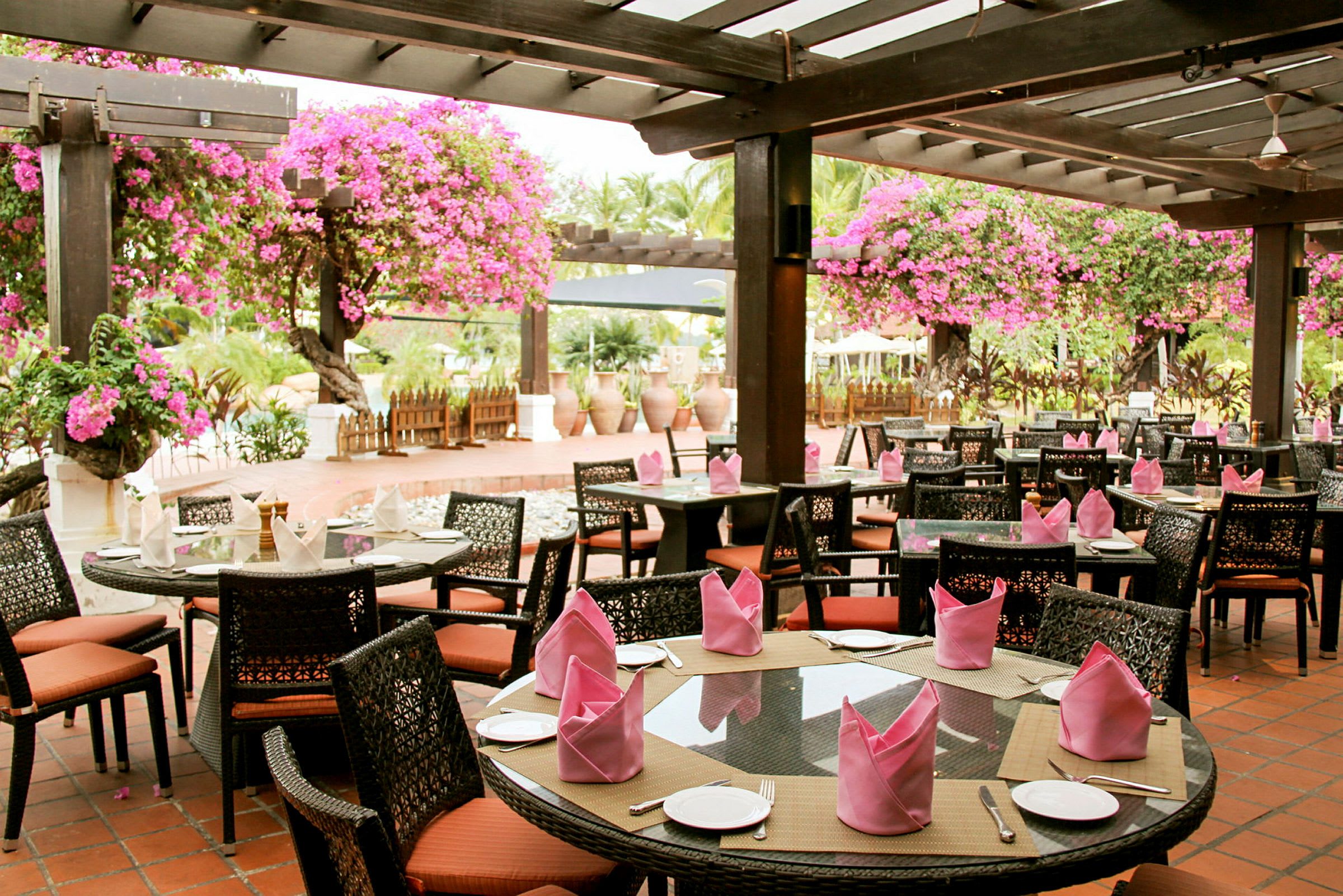 En utendørs restaurant med bord og stoler under en pergola dekorert med rosa blomster. (AI-generert tekst)