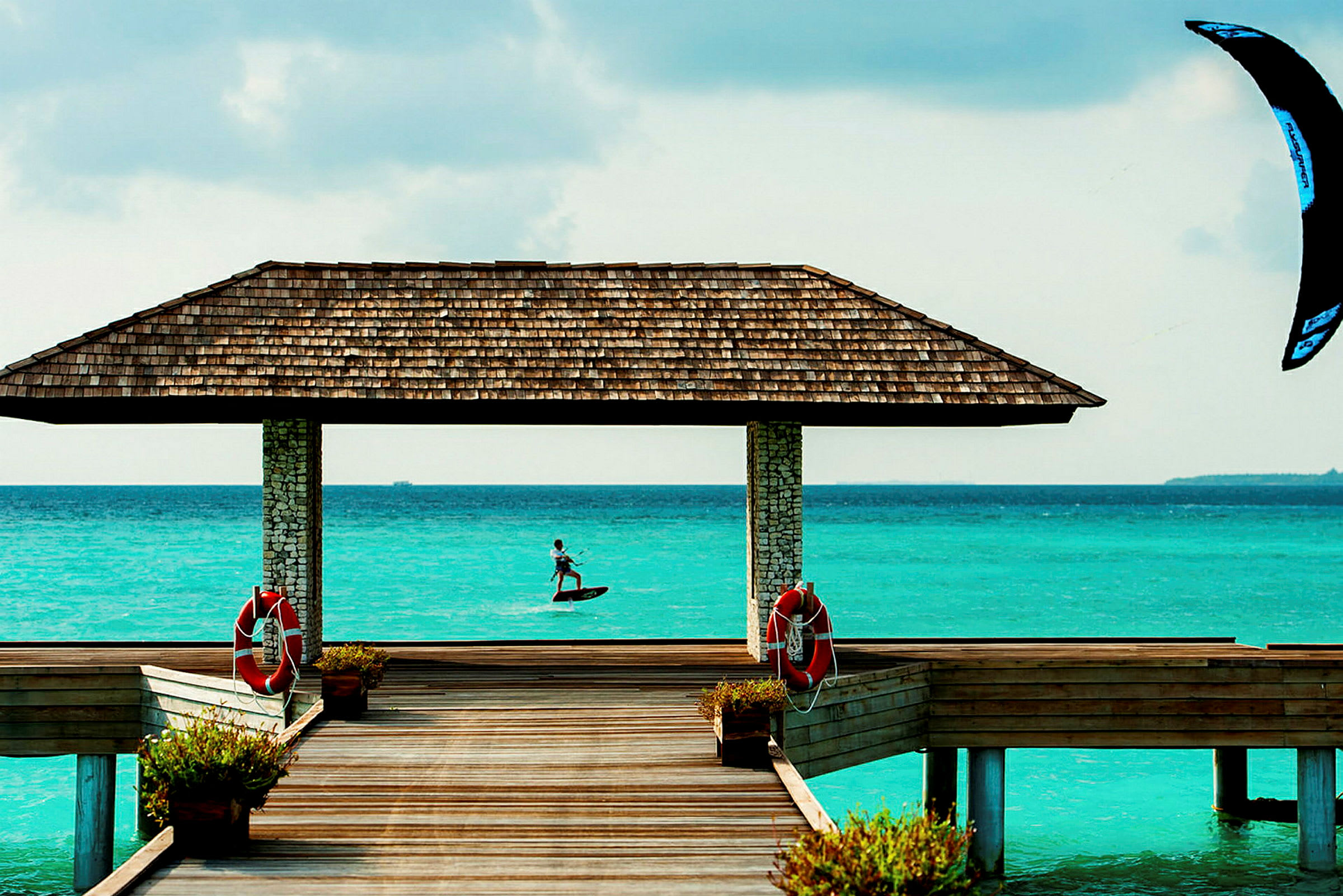 En trepier fører til et takoverheng ovenfor turkis vann, og en person kite-surfer i bakgrunnen. (AI-generert tekst)