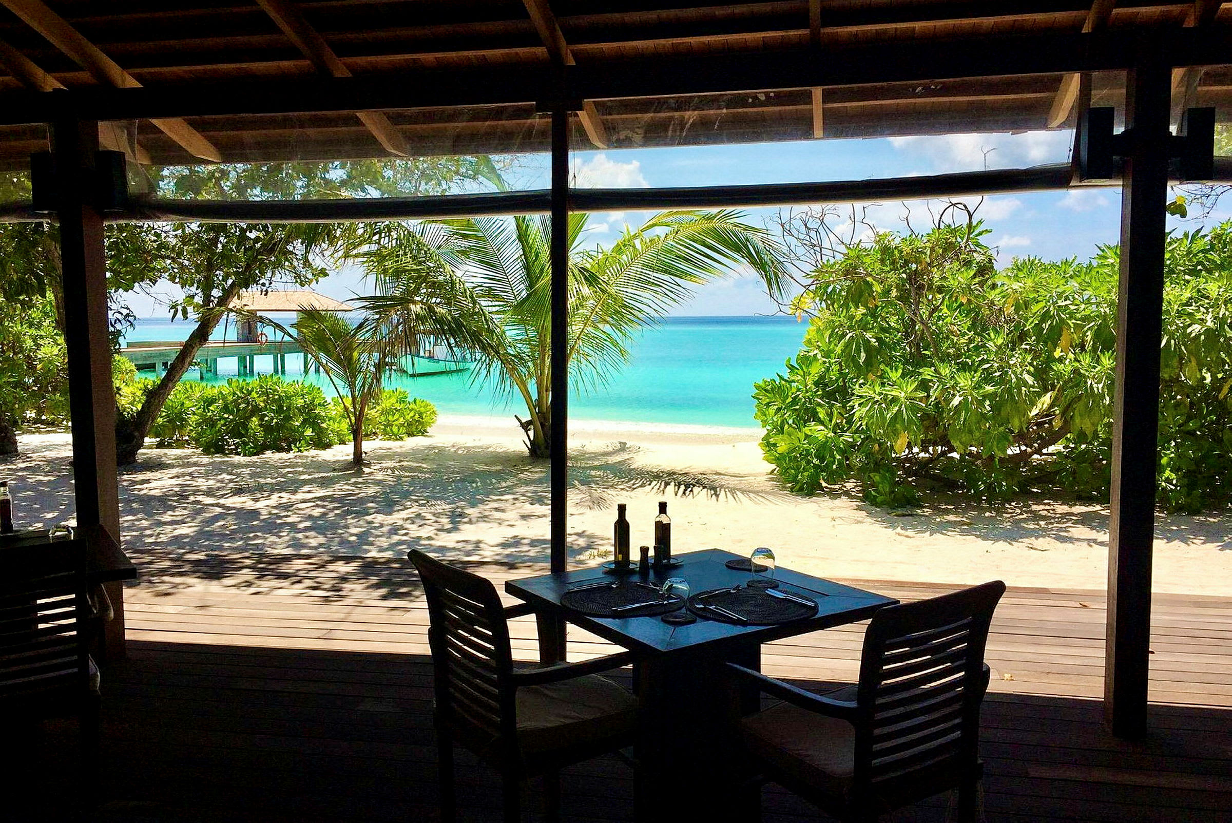 Utsikt fra en restaurant mot en tropisk strand med turkis hav og frodig vegetasjon. (AI-generert tekst)