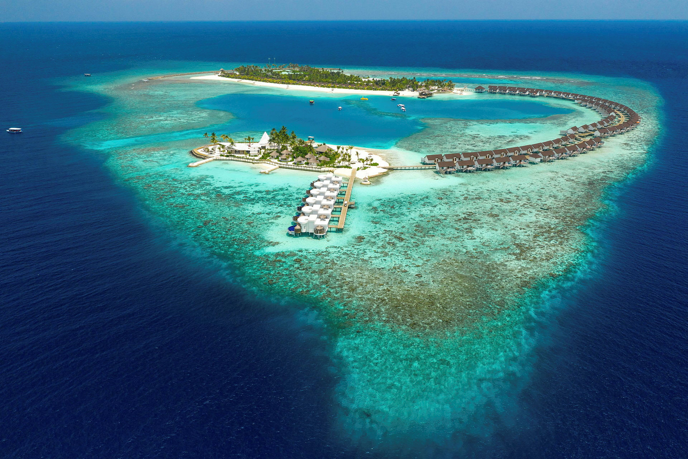 Flyfoto av en tropisk øy med overvannsbungalower og blå lagune omgitt av korallrev. (AI-generert tekst)