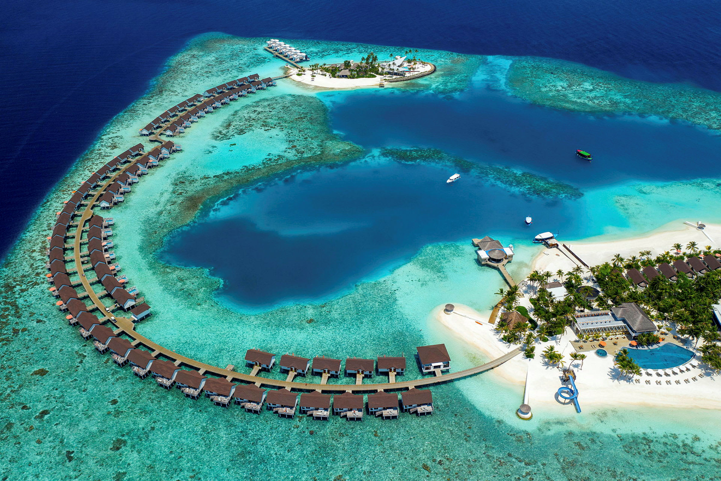 Et flyfoto av en tropisk ferie resort med bungalower på vannet og en sentral øy omgitt av turkis hav. (AI-generert tekst)