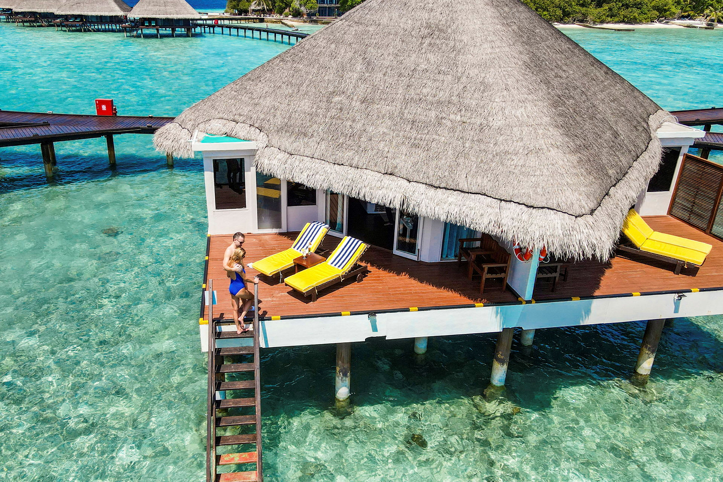 Bungalow på påler over klart turkis vann med solterrasse og strandstoler. (AI-generert tekst)