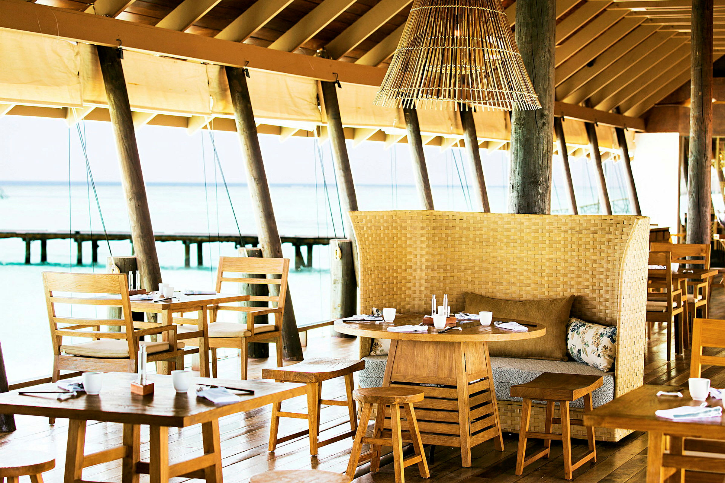 En restaurant med trebord og stoler samt en flettet benk med puter, plassert ved sjøen. (AI-generert tekst)