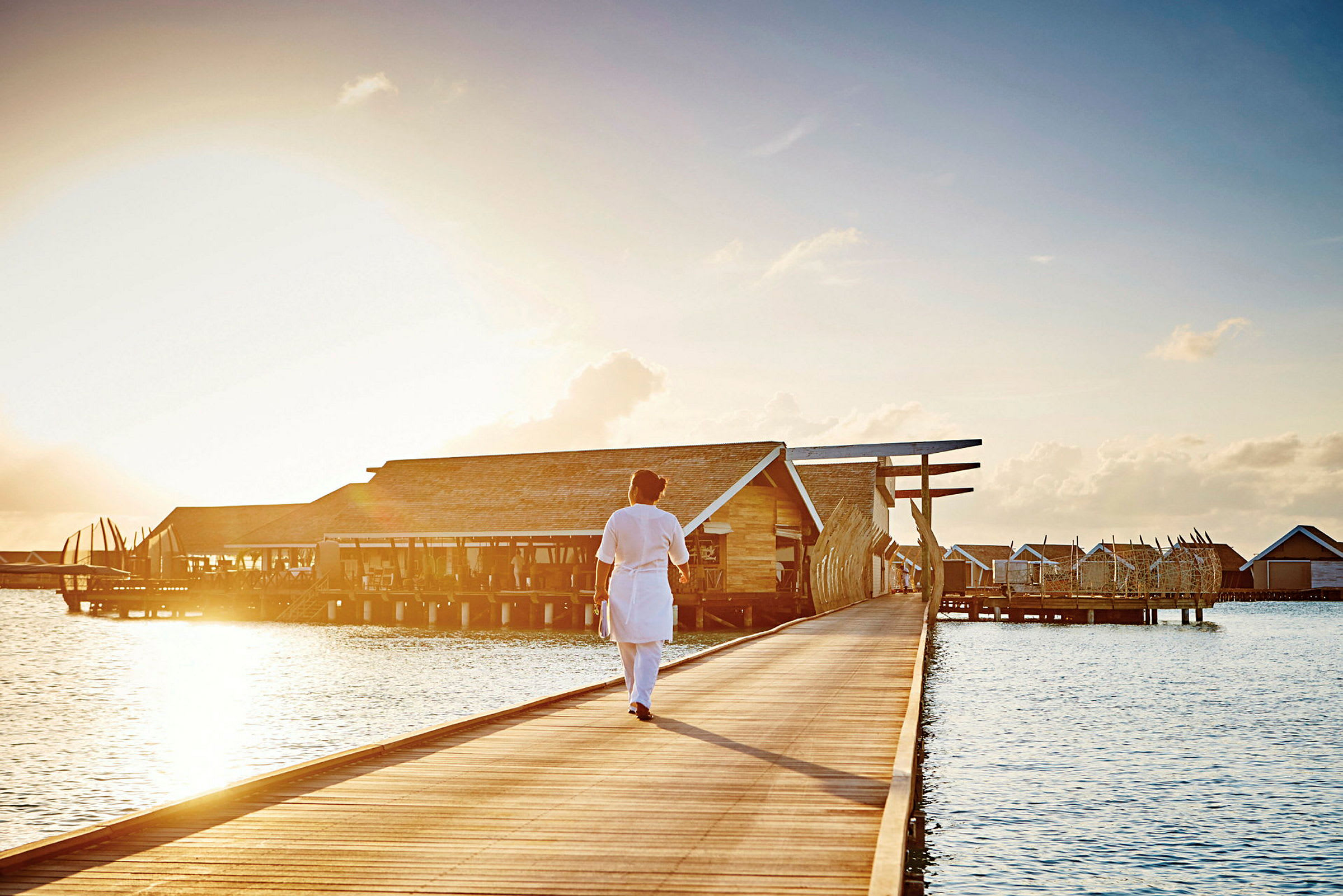 Person som går på en brygge mot strandhus over vannet ved solnedgang. (AI-generert tekst)