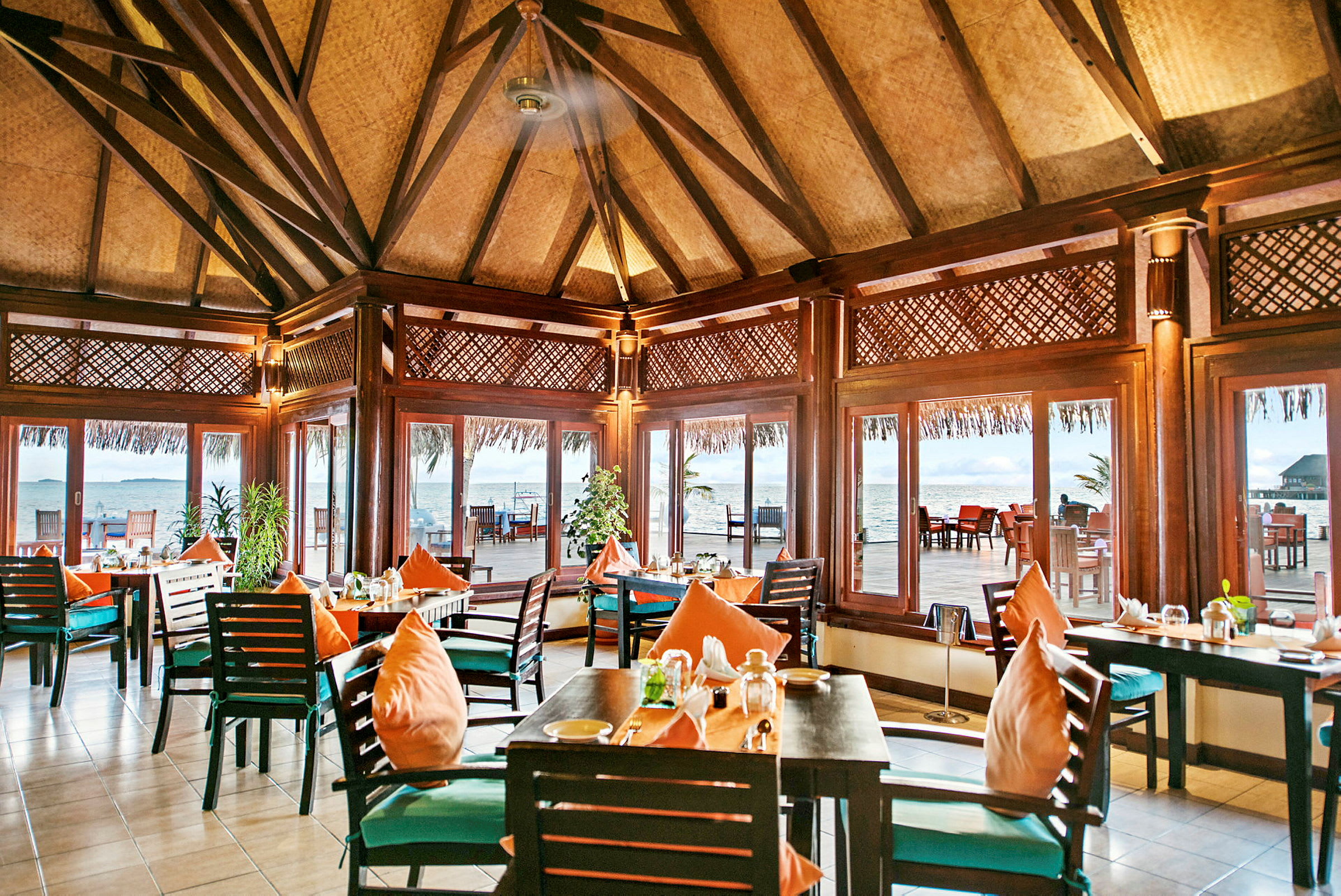 En restaurant med treinteriør og bord med oransje puter, omgitt av store vinduer med utsikt mot havet. (AI-generert tekst)