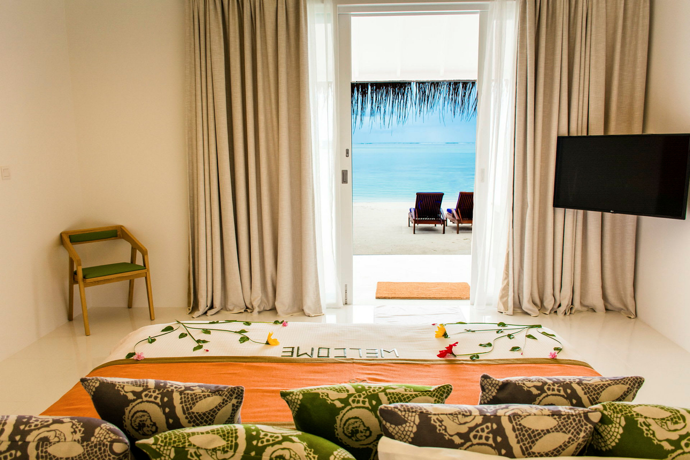 Utsikt fra hotellrom med dekorert seng, ryggstølstol og TV, mot strand og hav gjennom glassdør. (AI-generert tekst)