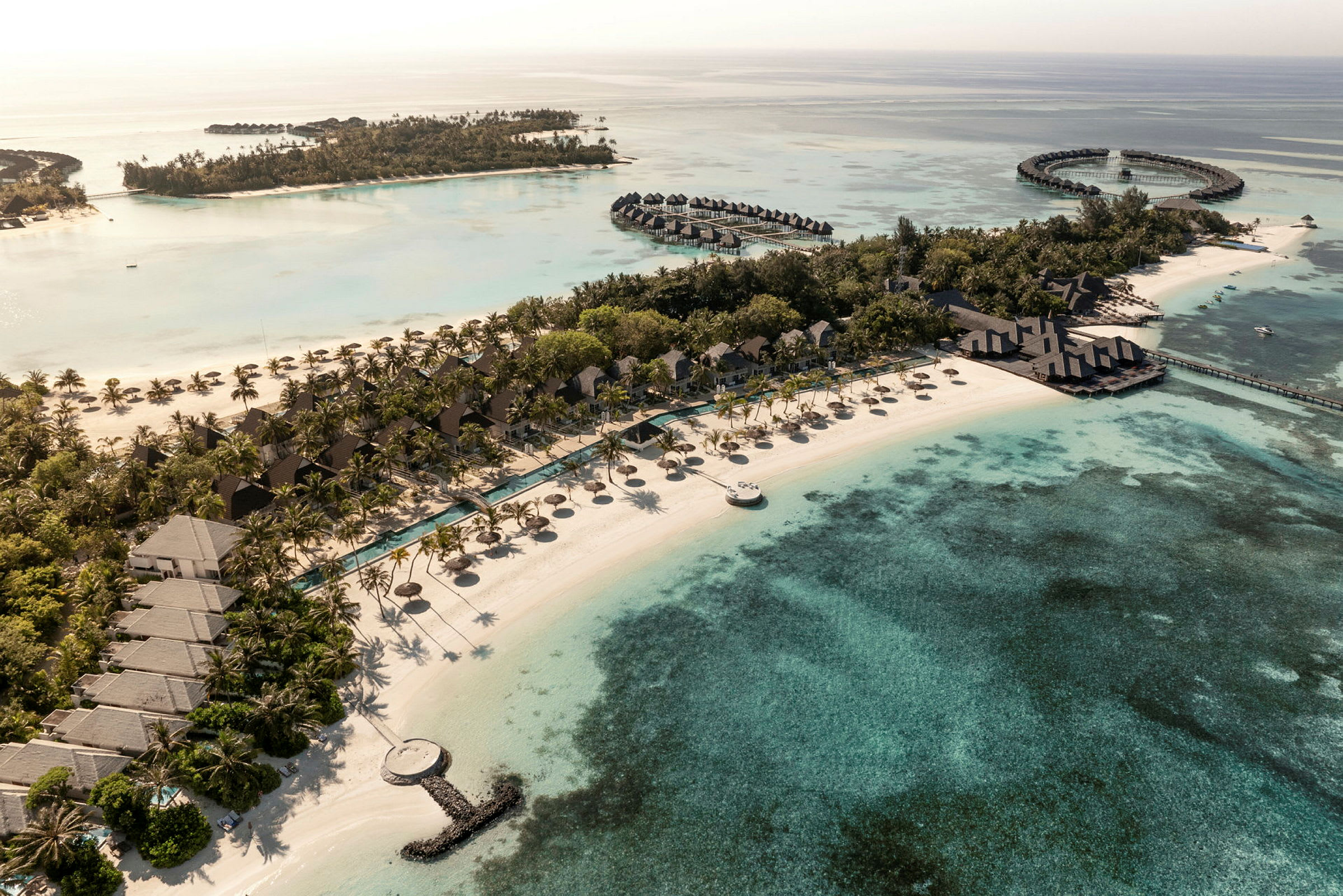 Flyfoto av en tropisk resort ved stranden med bungalower, bassenger og omkringliggende klarblått hav. (AI-generert tekst)