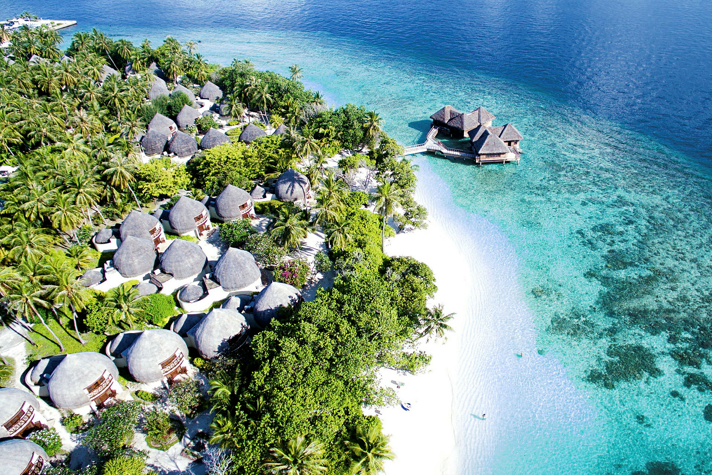 Flyfotografi av en tropisk kystlinje med bungalower blant palmer og en strand med turkist vann. (AI-generert tekst)