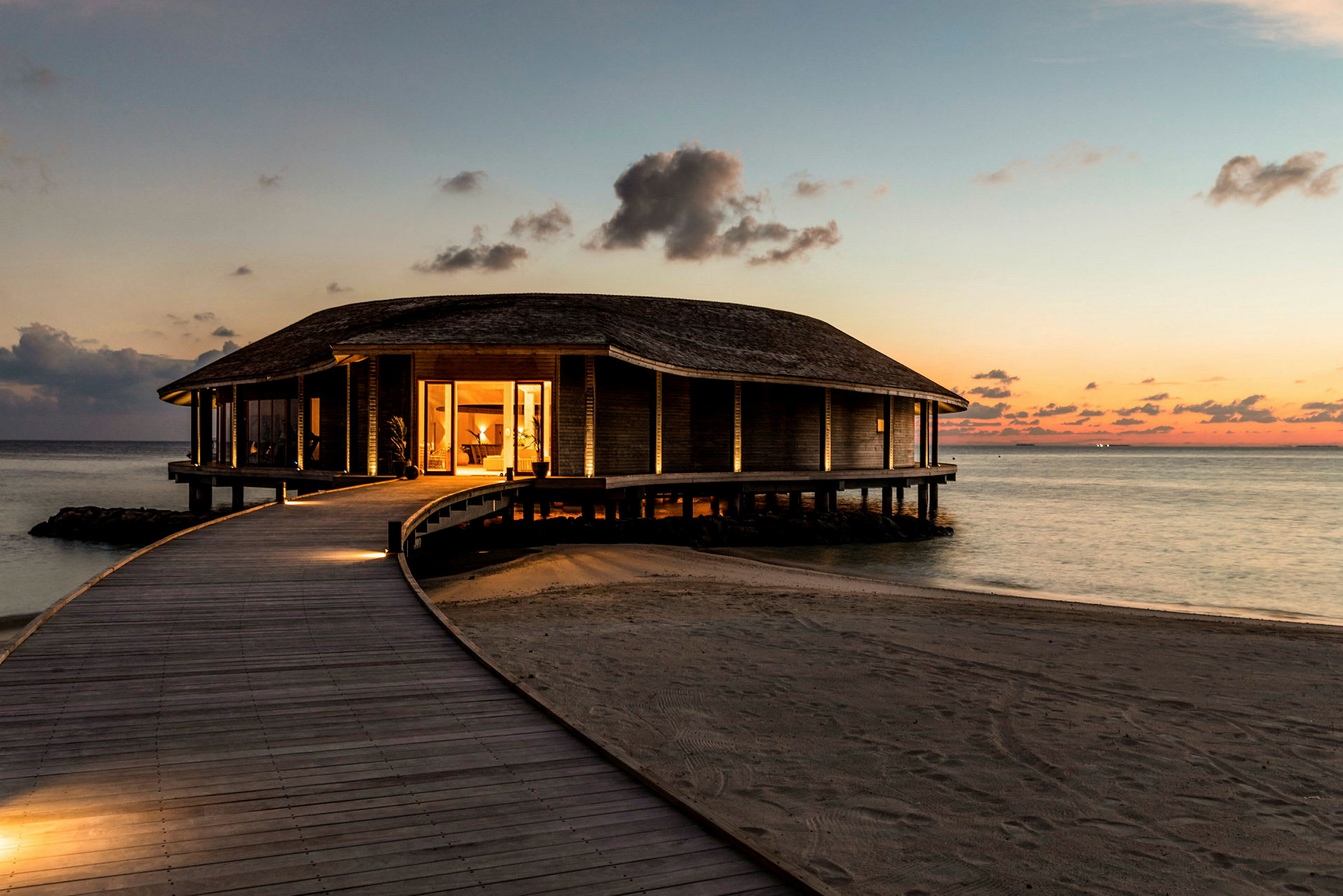 Et opplyst hus på påler ved havet med en tresti som fører frem til det, omgitt av sandstrand ved solnedgang. (AI-generert tekst)