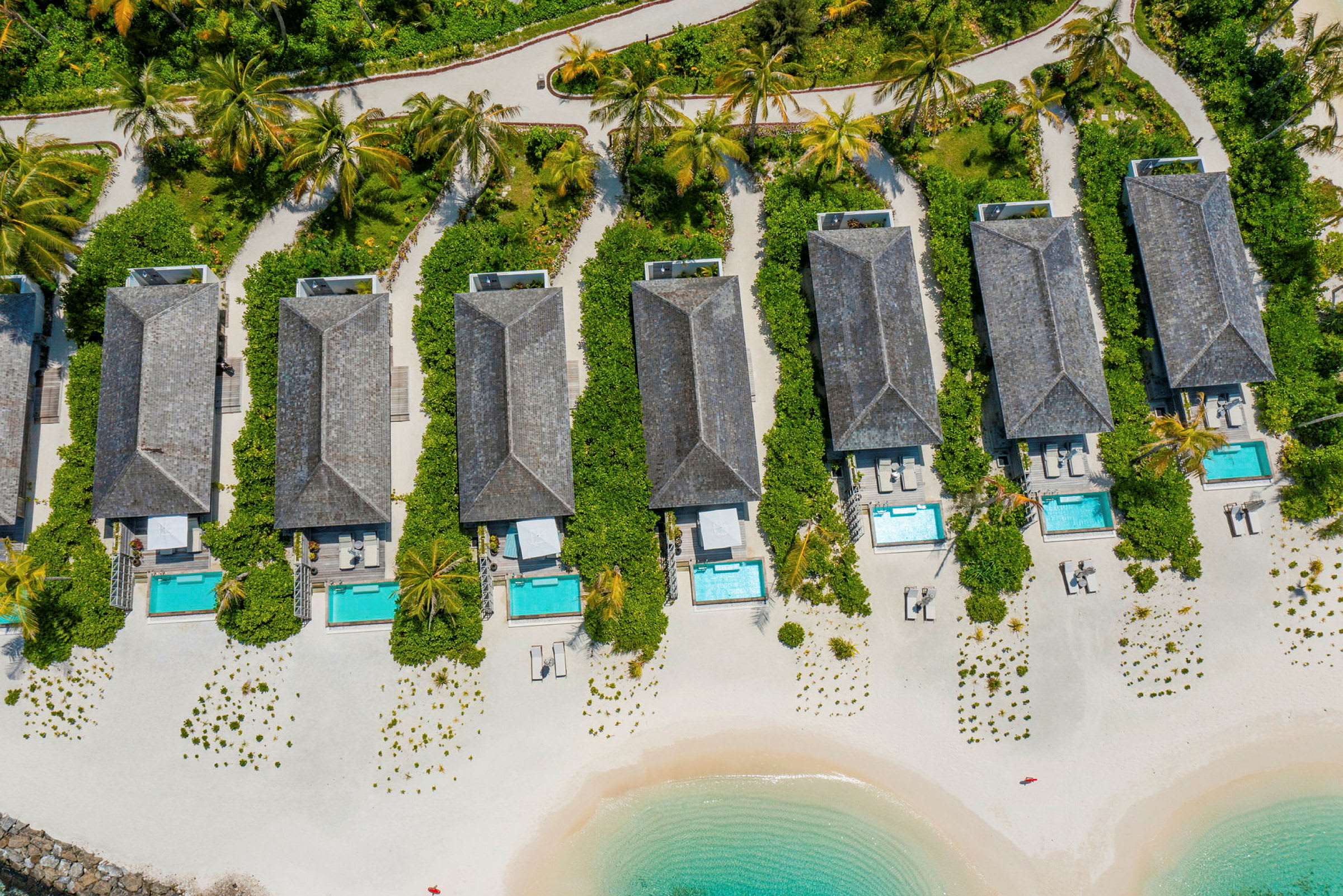 Flyfoto av strandvillaer med private bassenger og hager på en sandstrand ved havet. (AI-generert tekst)