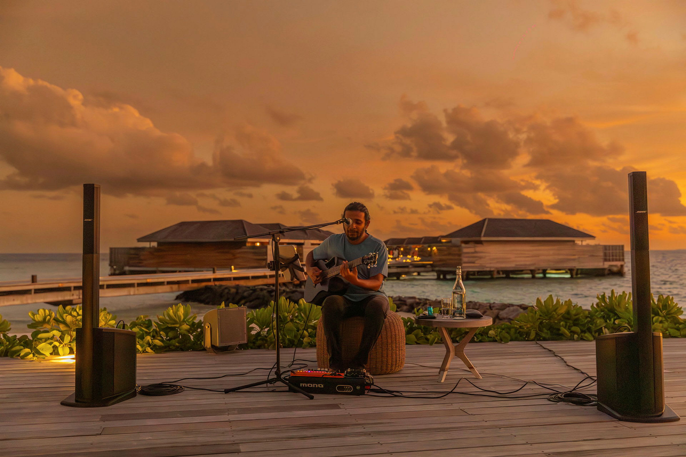 En person spiller gitar mens solen går ned ved en strand med vannbungalower. (AI-generert tekst)