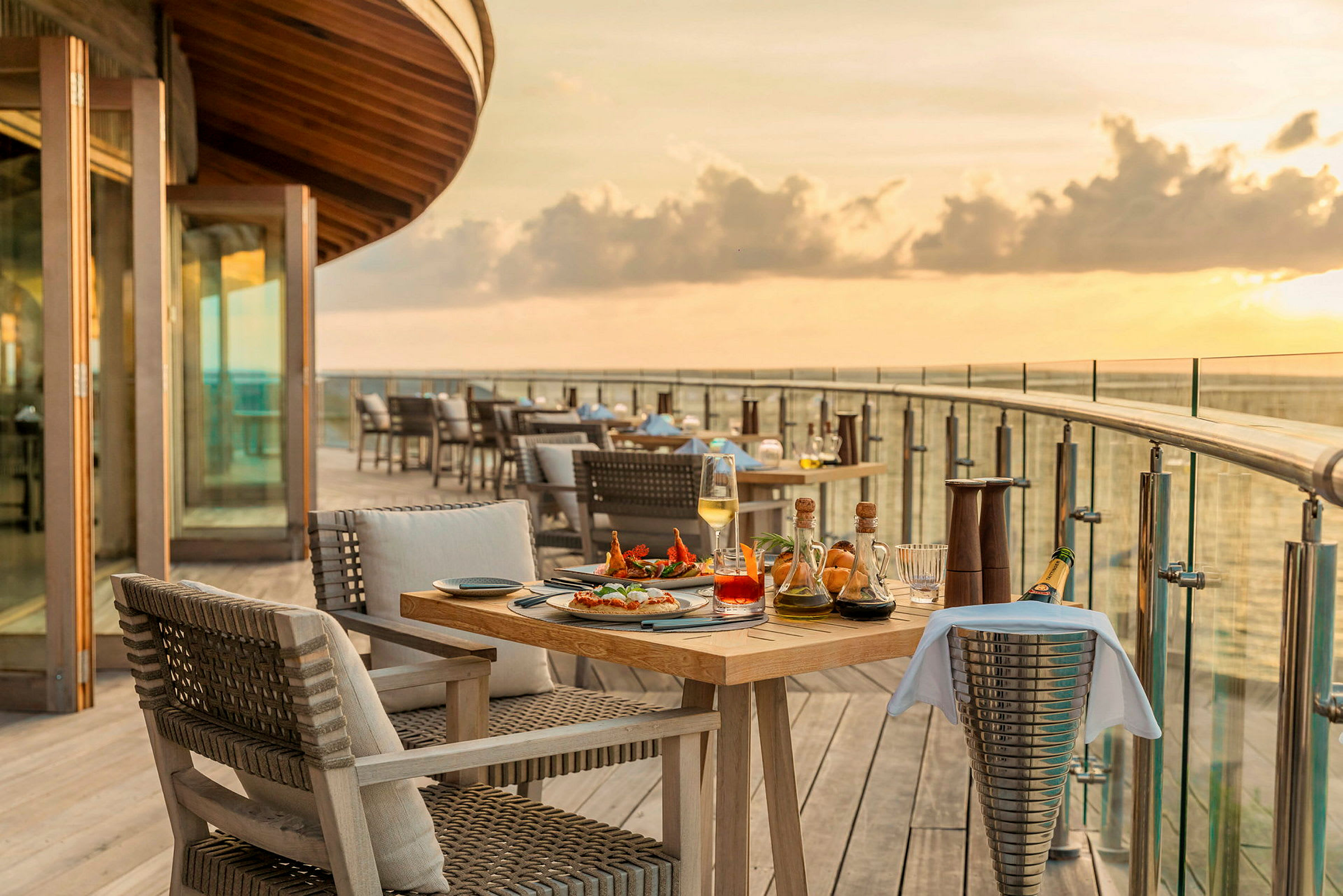 Spisebord med mat og drikke på en treterrasse med havutsikt ved solnedgang. (AI-generert tekst)