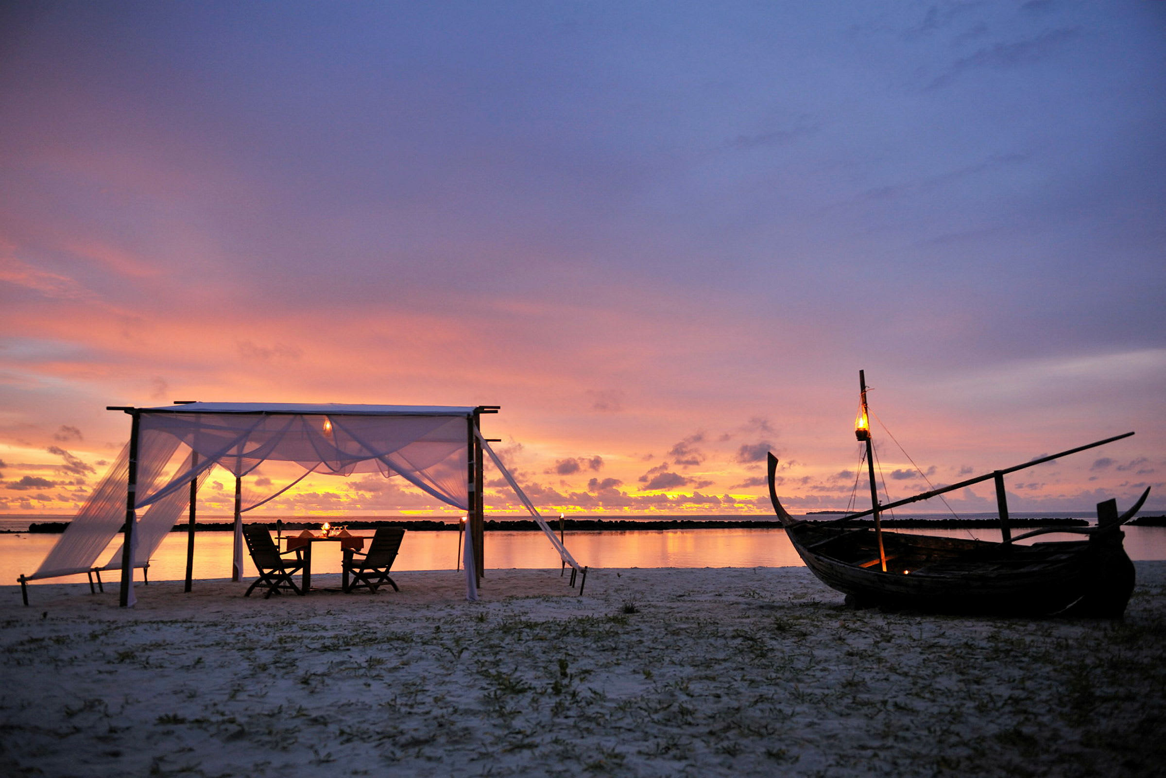 En strand med en liten baldakin og stoler ved solnedgang, med en trebåt og lykter. (AI-generert tekst)