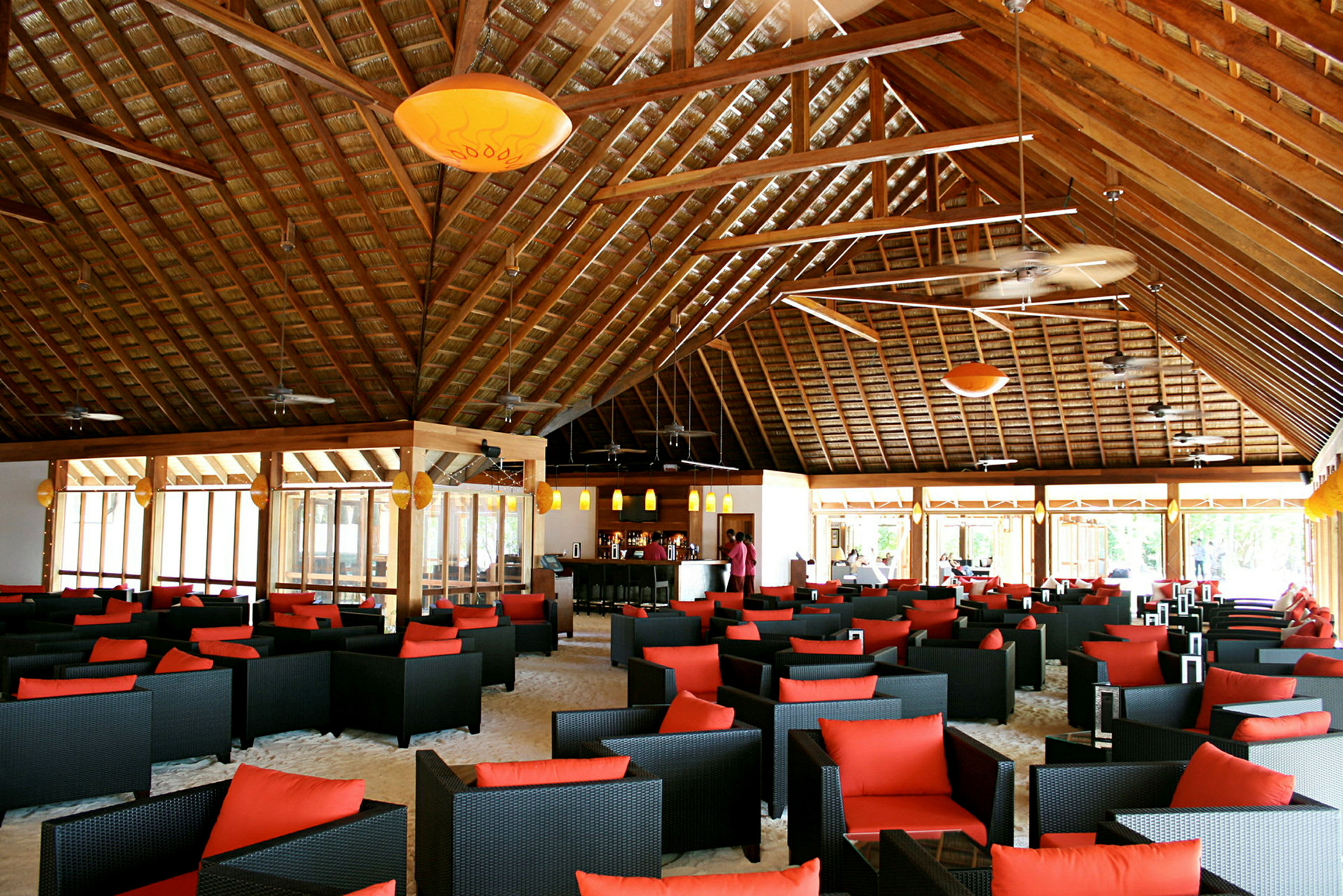 Et interiør av en romslig restaurant eller lounge med svarte flettede stoler med røde puter, bord plassert på sandgulv under et høyt trebjelketak. (AI-generert tekst)