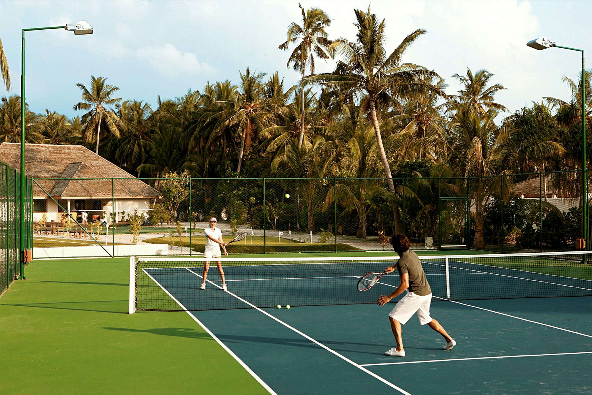 To personer spiller tennis på en utendørsbane omgitt av palmer. (AI-generert tekst)