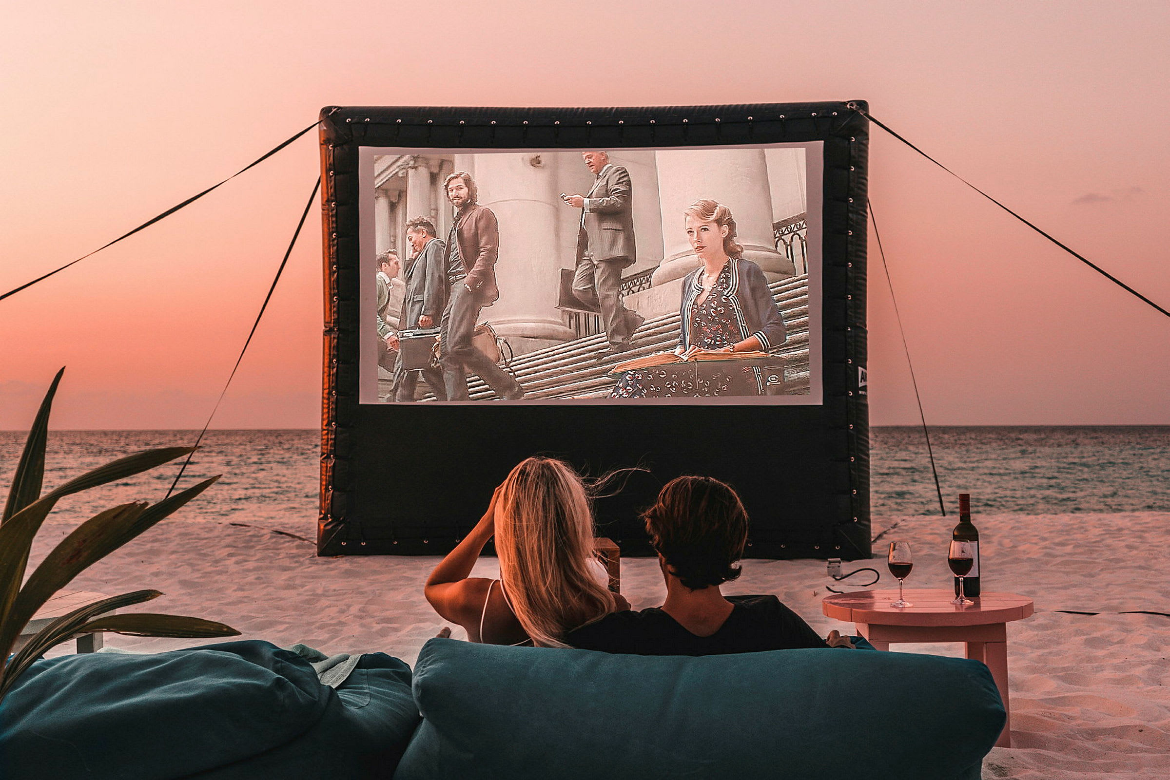 To personer sitter på stranden og ser på en film som vises på et utendørs lerret. (AI-generert tekst)