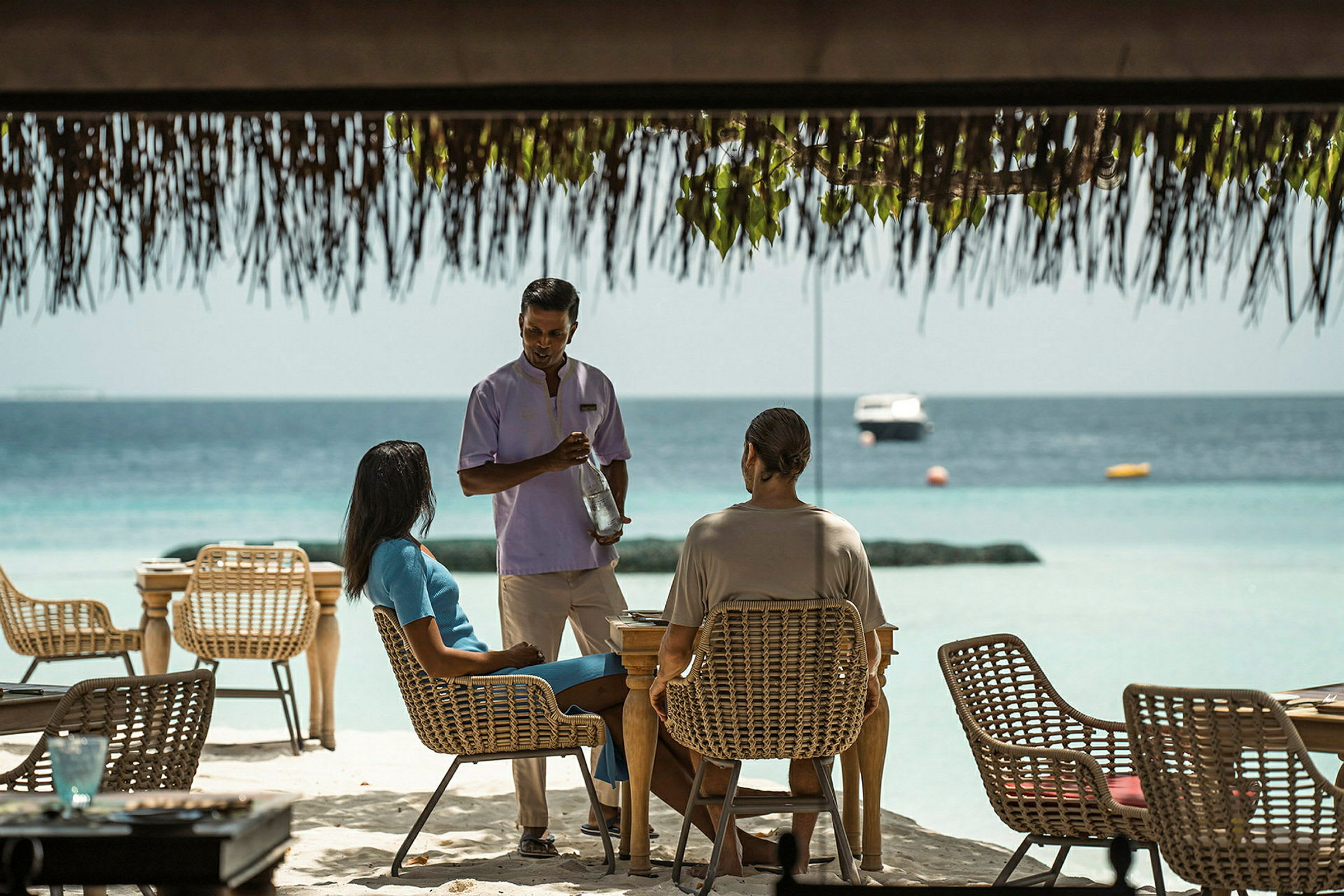 Servitør serverer gjester ved et bord på en utendørsrestaurant med utsikt over havet. (AI-generert tekst)