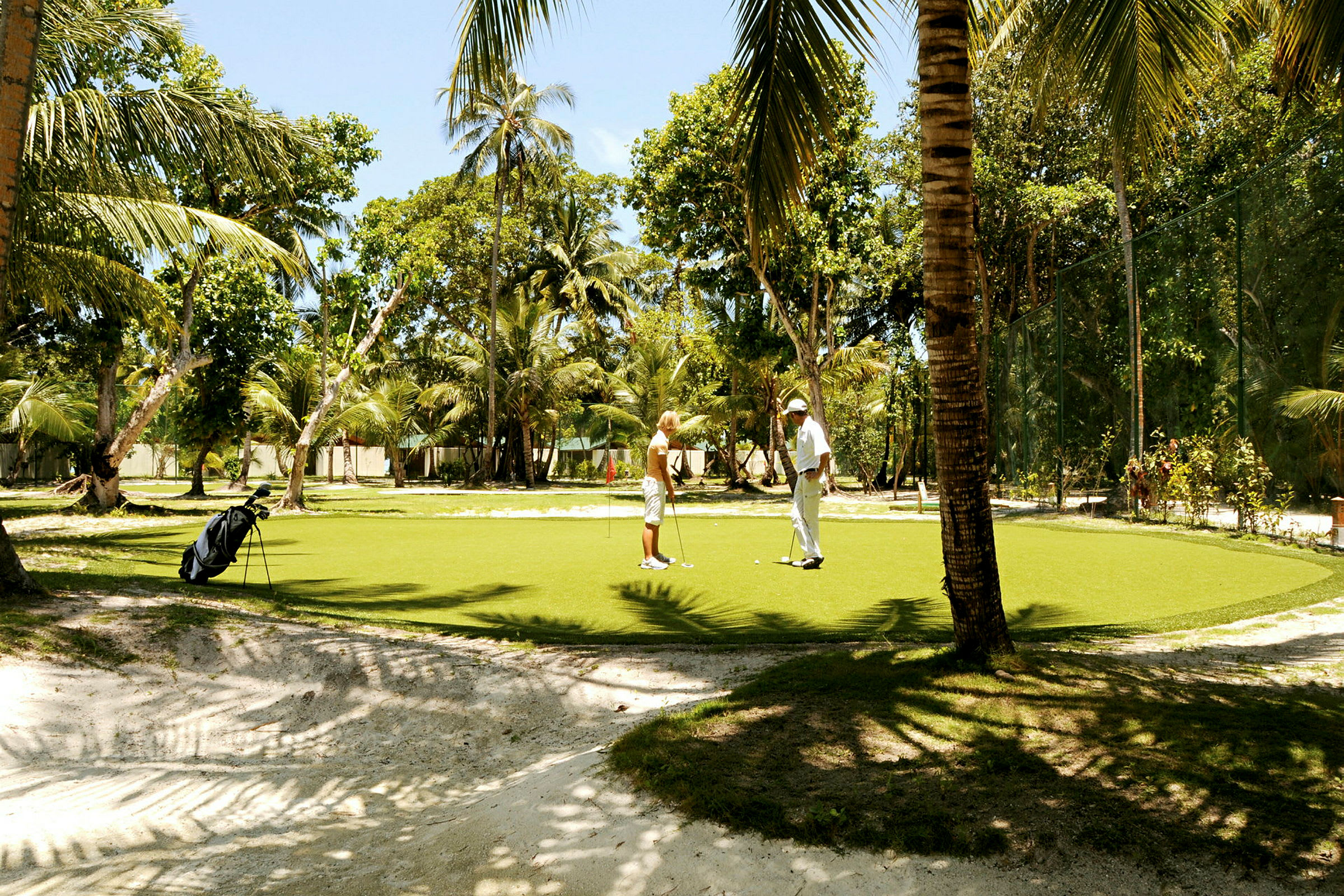 To personer spiller golf på en grønn puttinggreen omgitt av palmer. (AI-generert tekst)