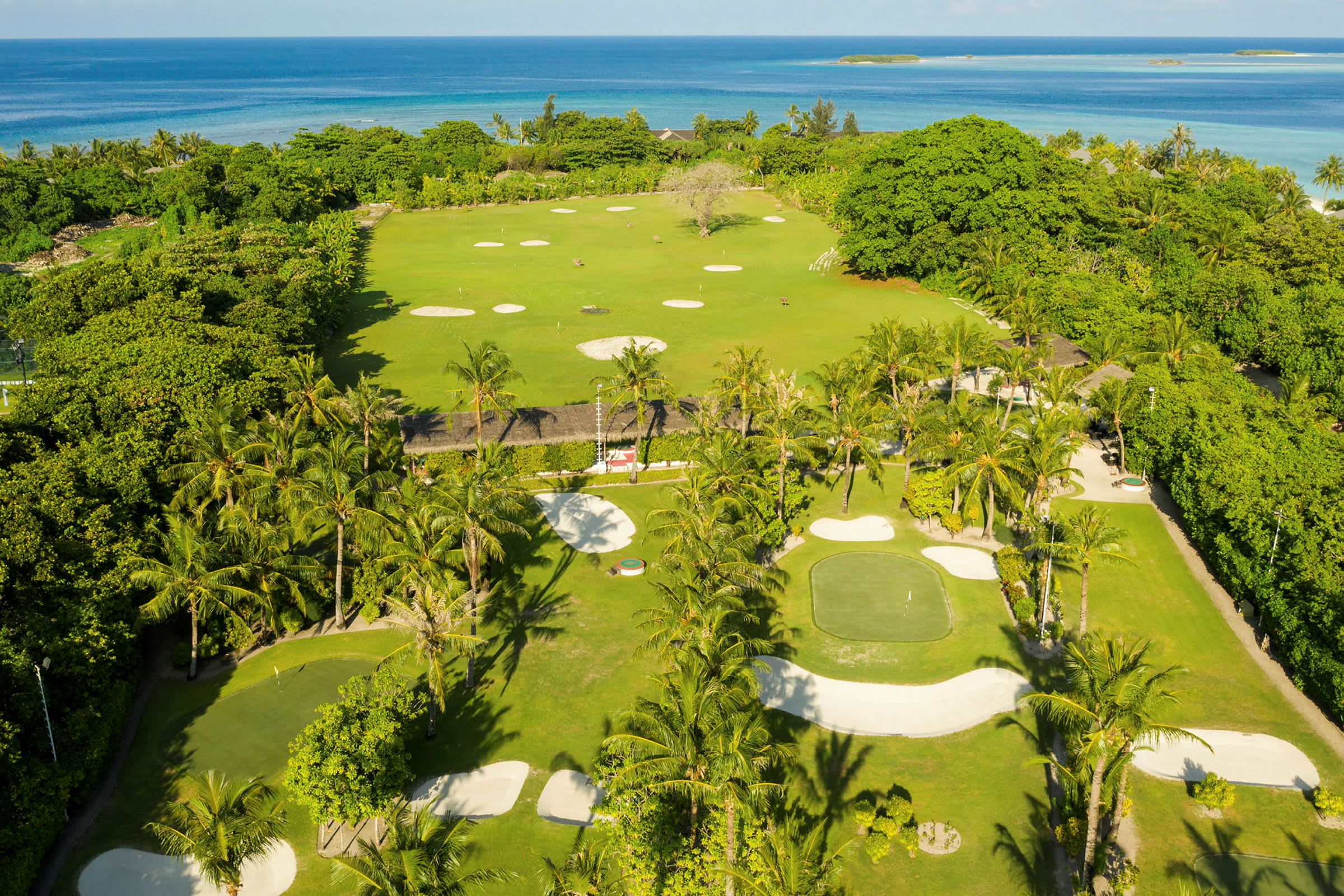 Utsikt over en golfbane med grønne områder, sandbunkere og palmer ved havet. (AI-generert tekst)