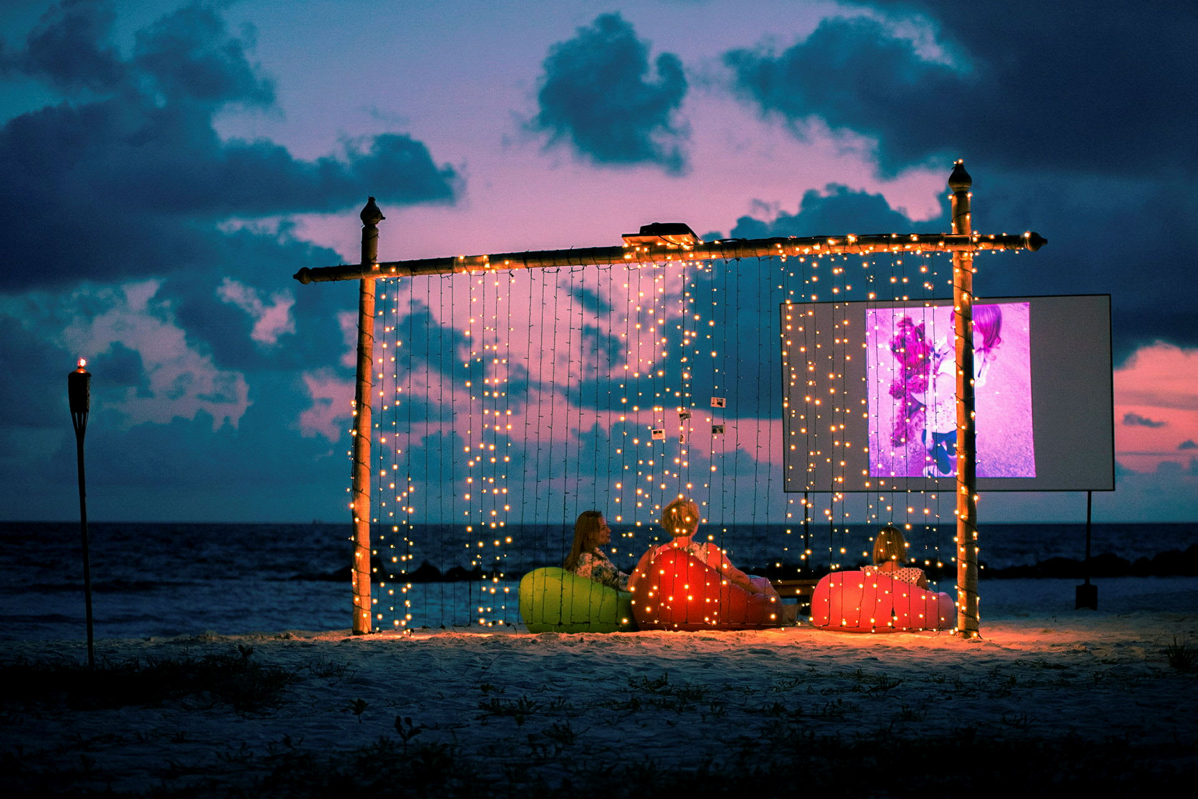Folk sitter ved en utendørs filmvisning på stranden under en rosa og blå skumring. (AI-generert tekst)