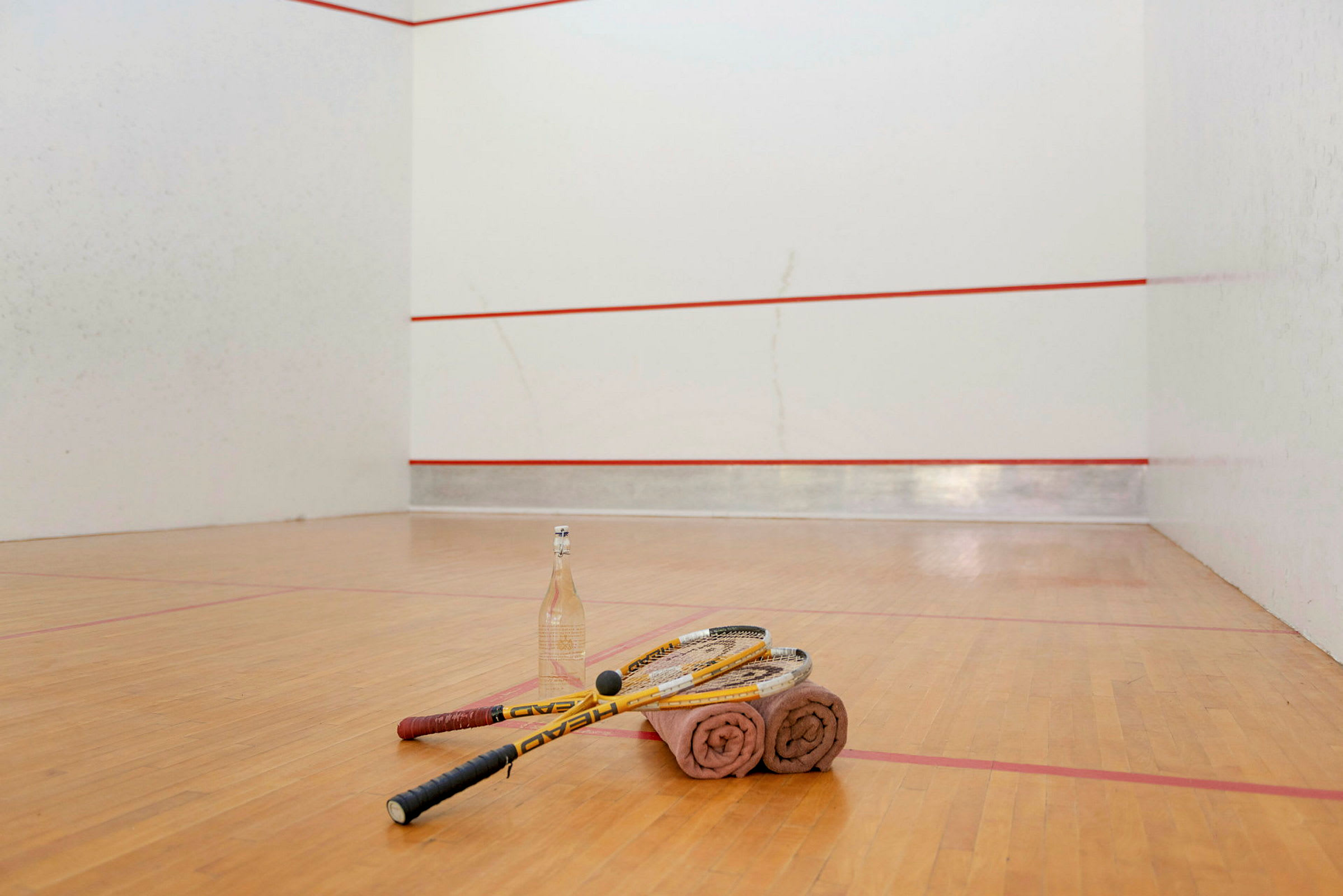 Squashracketer, håndklær og en vannflaske ligger på gulvet i en squashbane. (AI-generert tekst)