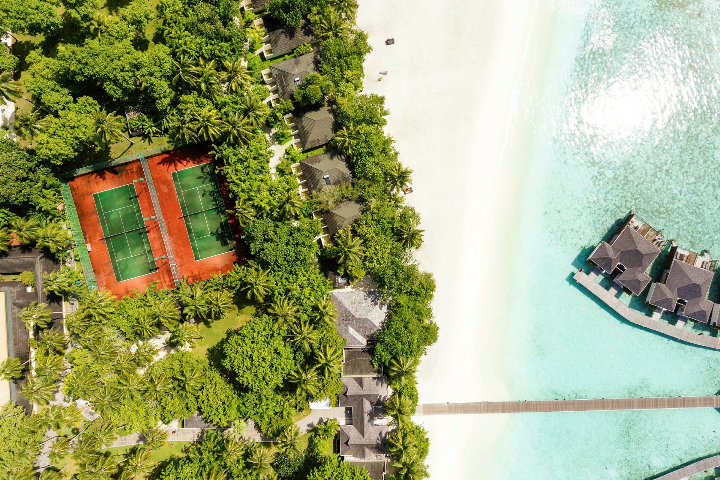 Flyfoto av en tropisk resort ved stranden med tennisbaner, villaer blant trær og overvannsbungalower. (AI-generert tekst)