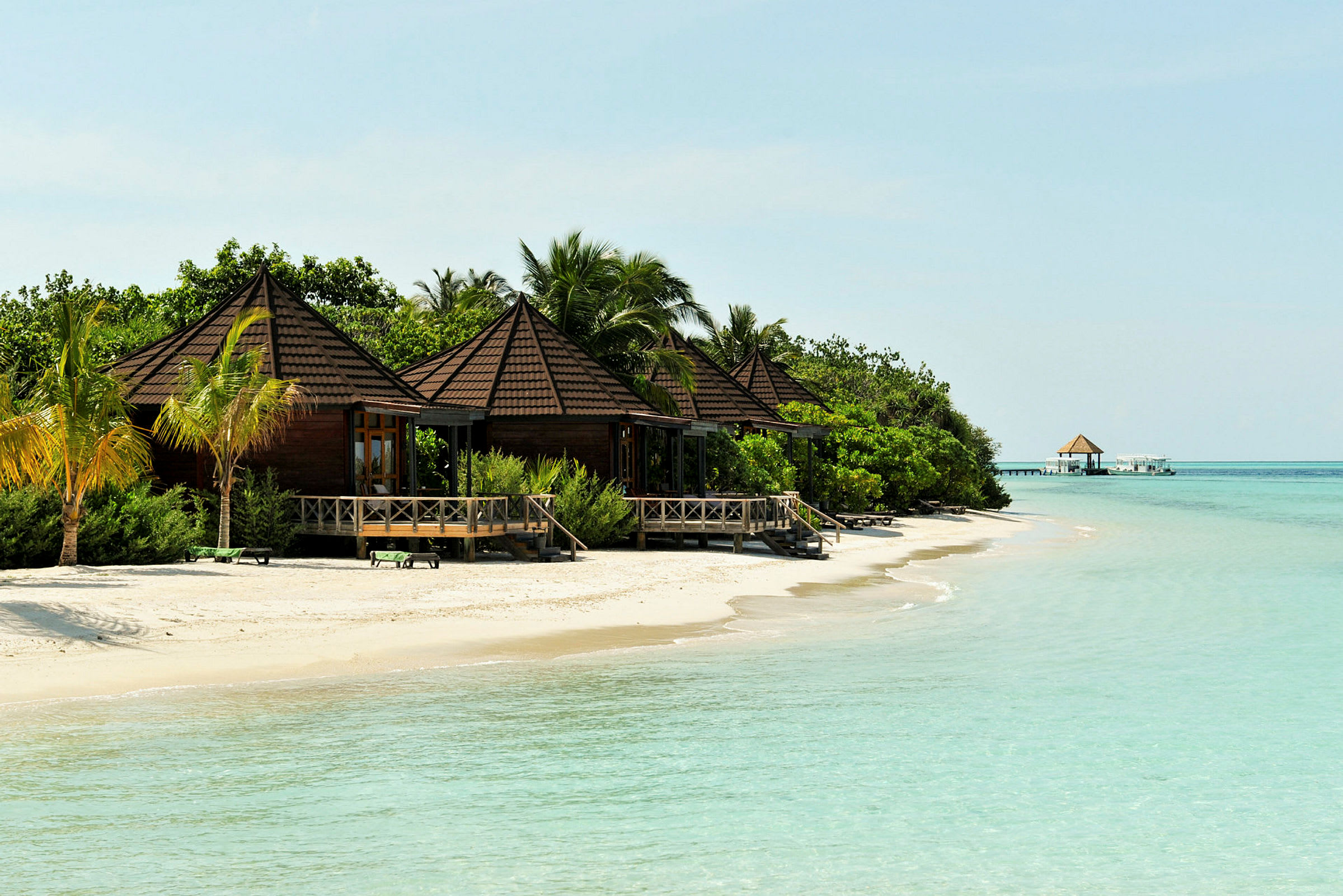 Flere trebungalower med høye tak ligger ved en sandstrand og grenser til klart, turkis vann. (AI-generert tekst)