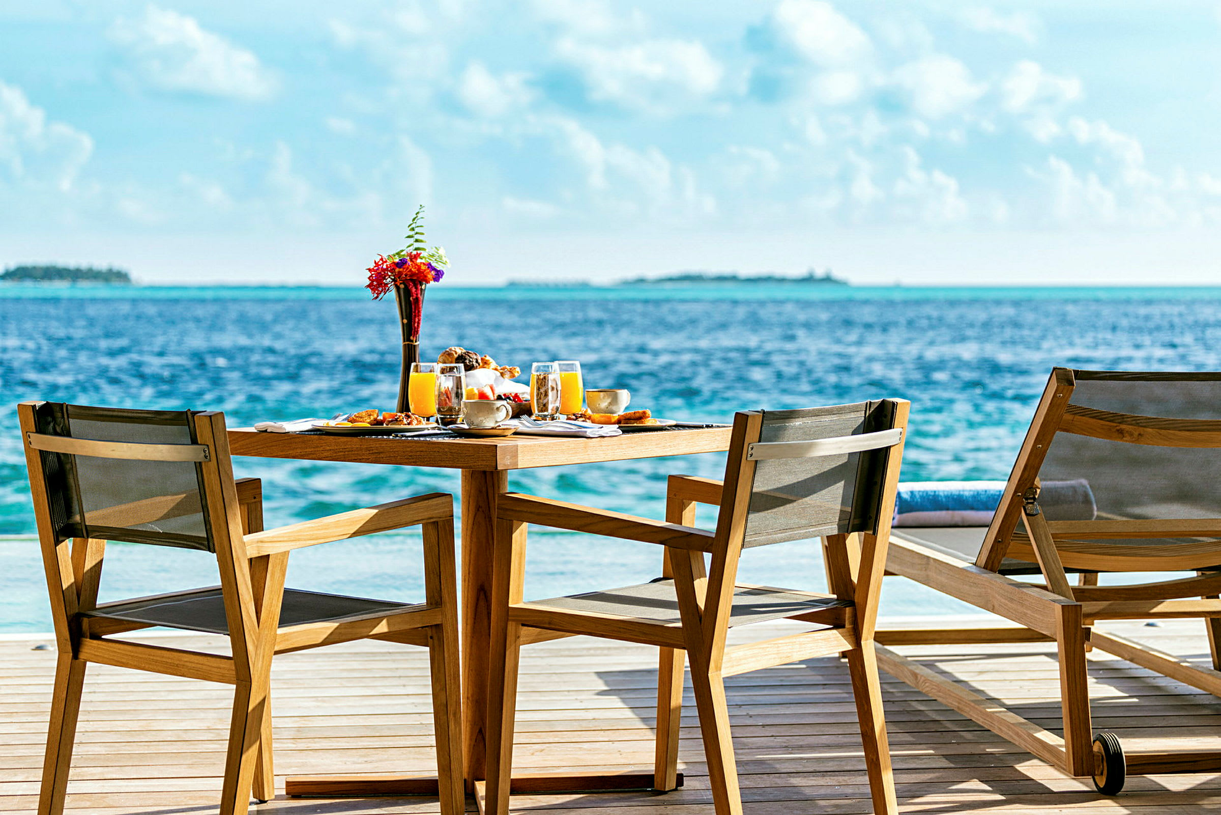 To hagestoler og et bord står på en treterrasse ved havet med en borddekking og utsikt over vannet. (AI-generert tekst)