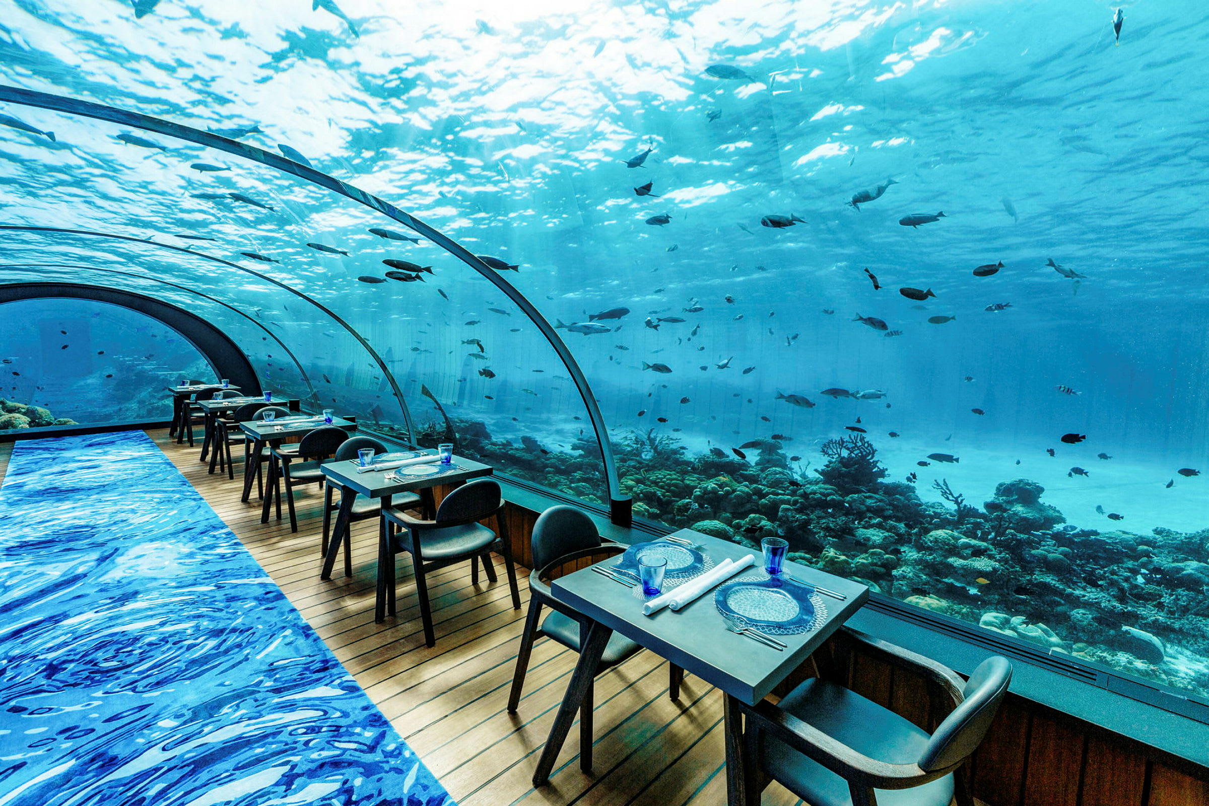 Undervannsområde med en restaurant hvor bord og stoler er plassert foran et stort, buet vindu med utsikt mot havet og fisk. (AI-generert tekst)