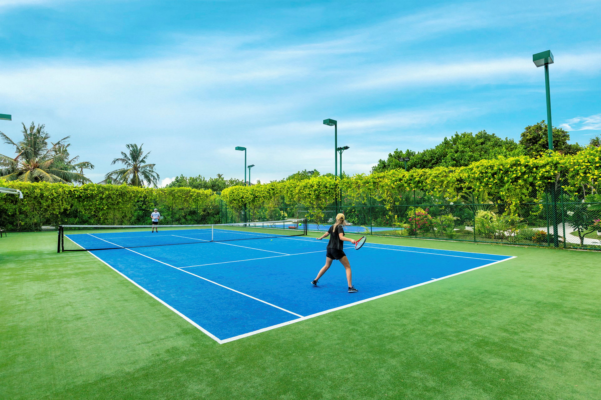 To personer spiller tennis på en blå tennisbane omgitt av grønt landskap. (AI-generert tekst)