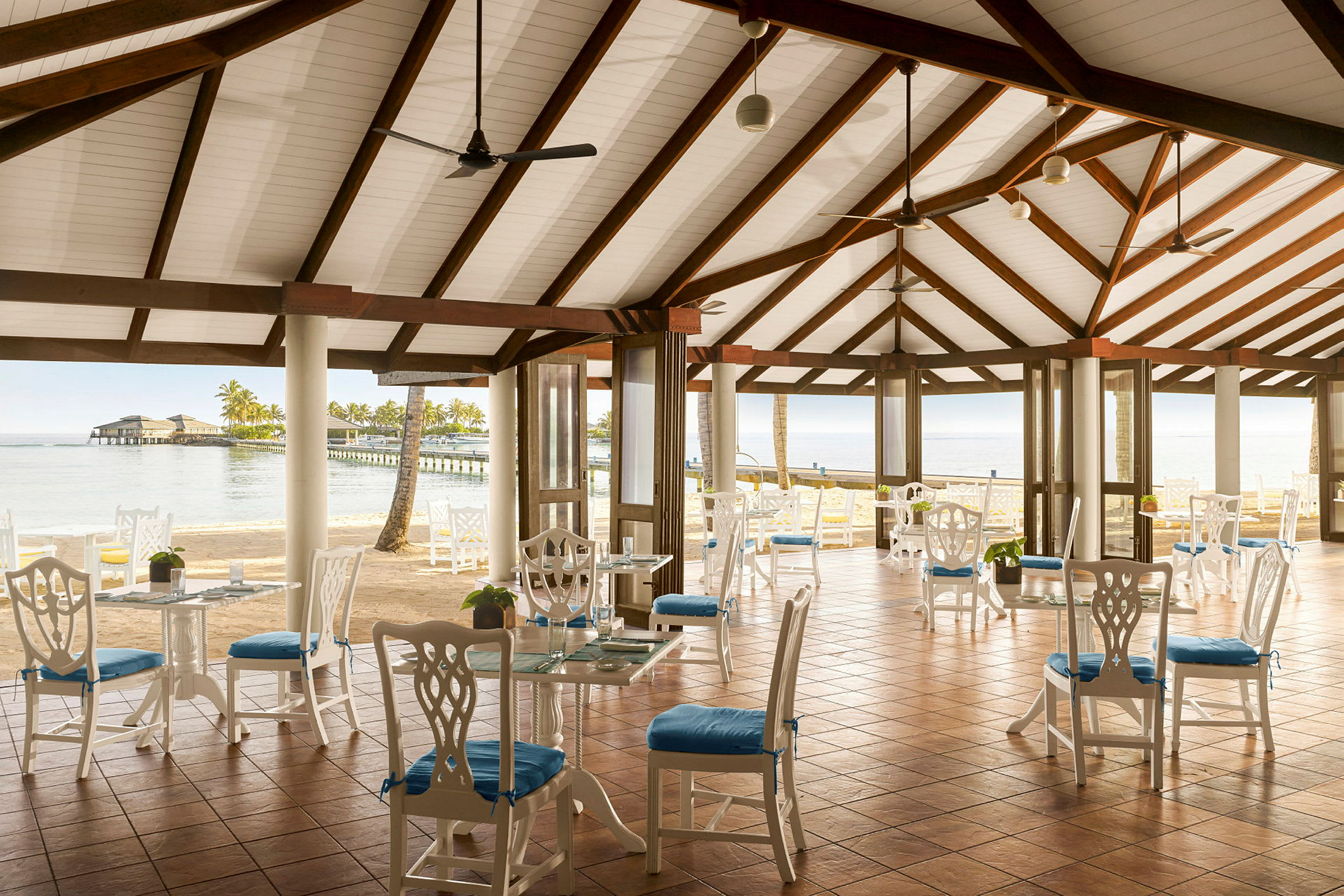 Bilde av en åpen restaurant med hvitmalte møbler og blå sitteputer, med utsikt mot havet og en brygge. (AI-generert tekst)