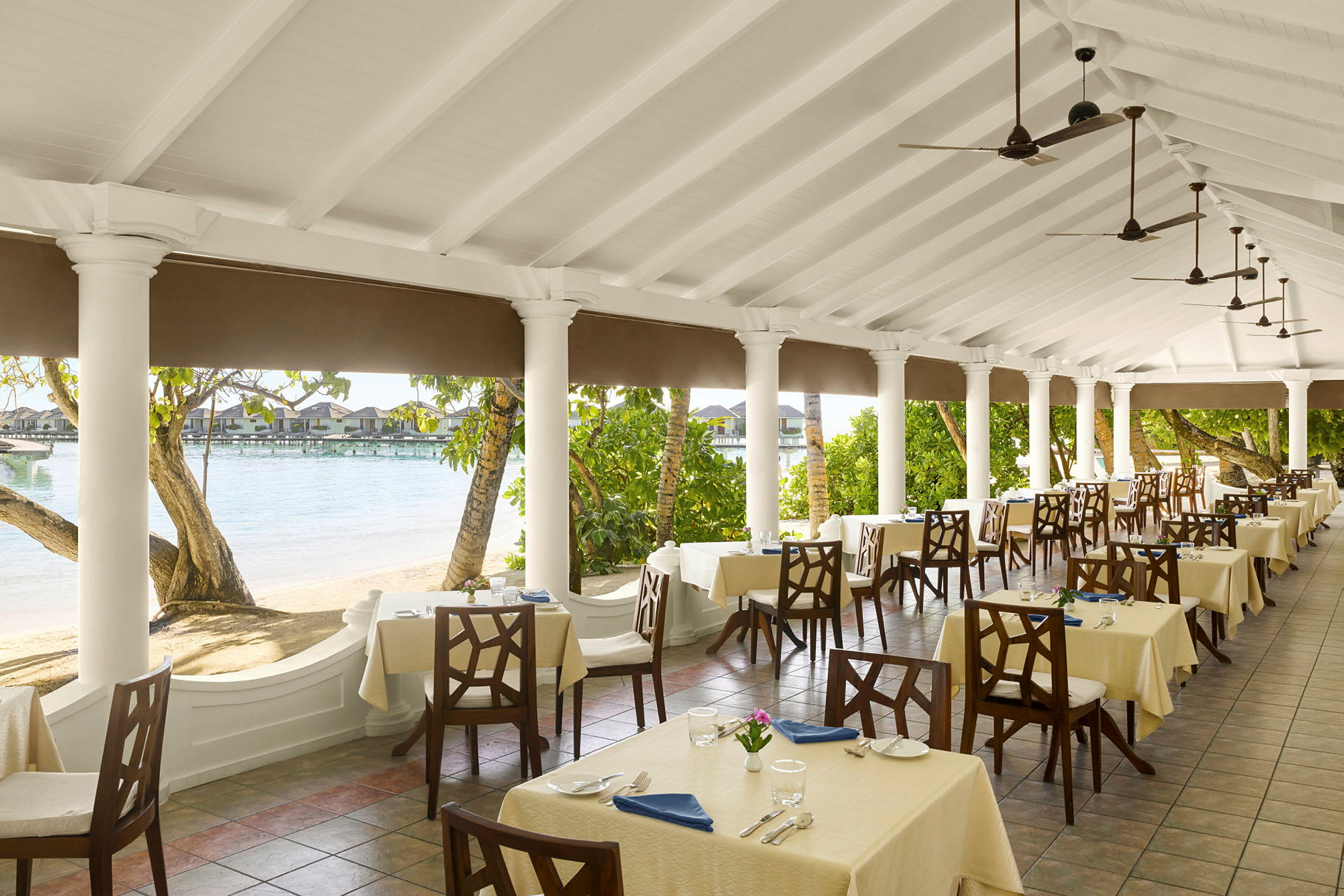 En restaurantterrasse med dekkede bord, omgitt av søyler og utsikt over stranden og vannet. (AI-generert tekst)